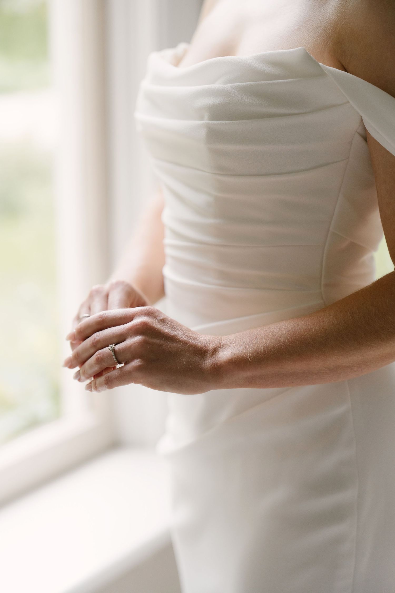 Close-up of bride in off-shoulder wedding dress showing engagement ring by window.