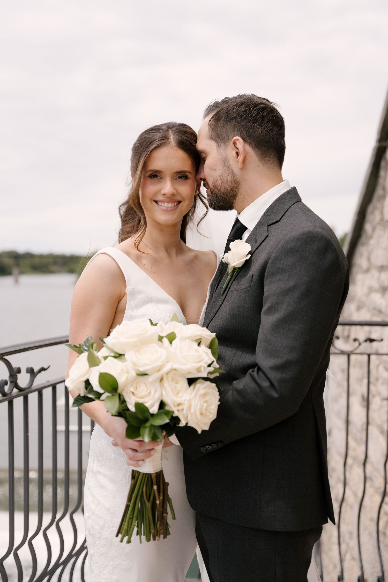 Smiling bride holding white rose bouquet as groom whispers to her on a waterfront balcony.