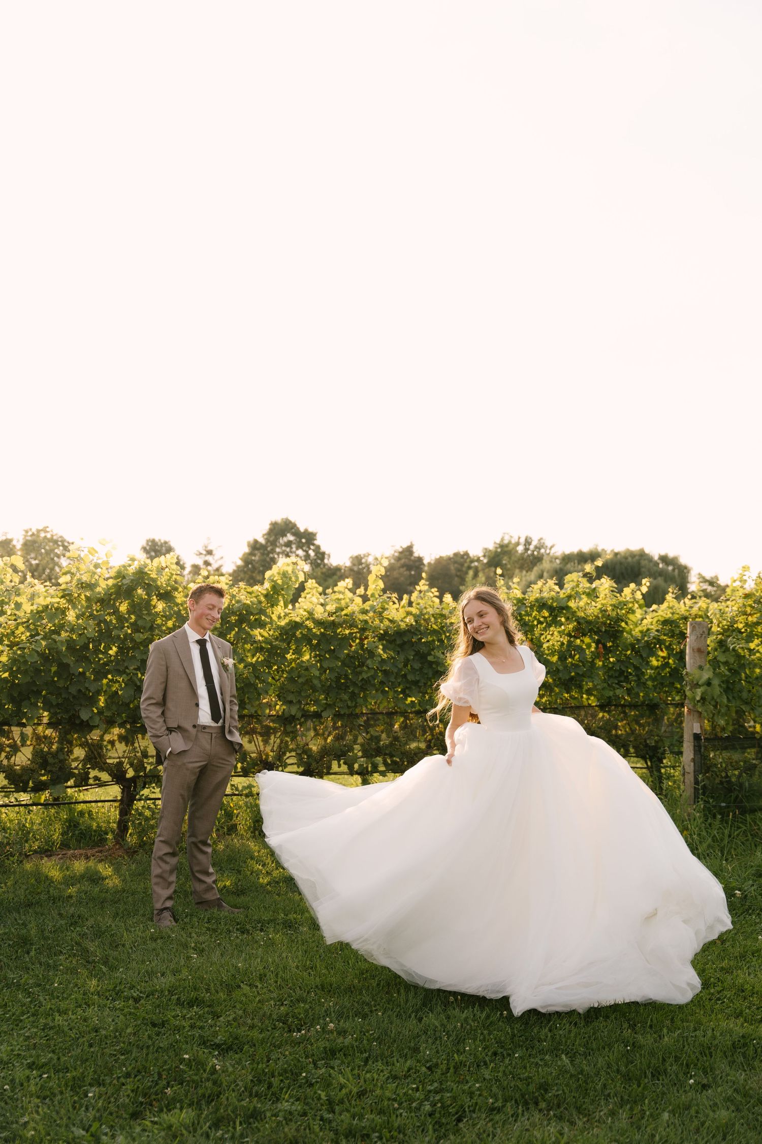 Bride twirling her white ball gown skirt in a Niagara-on-the-lake vineyard while groom watches adoringly at sunset.