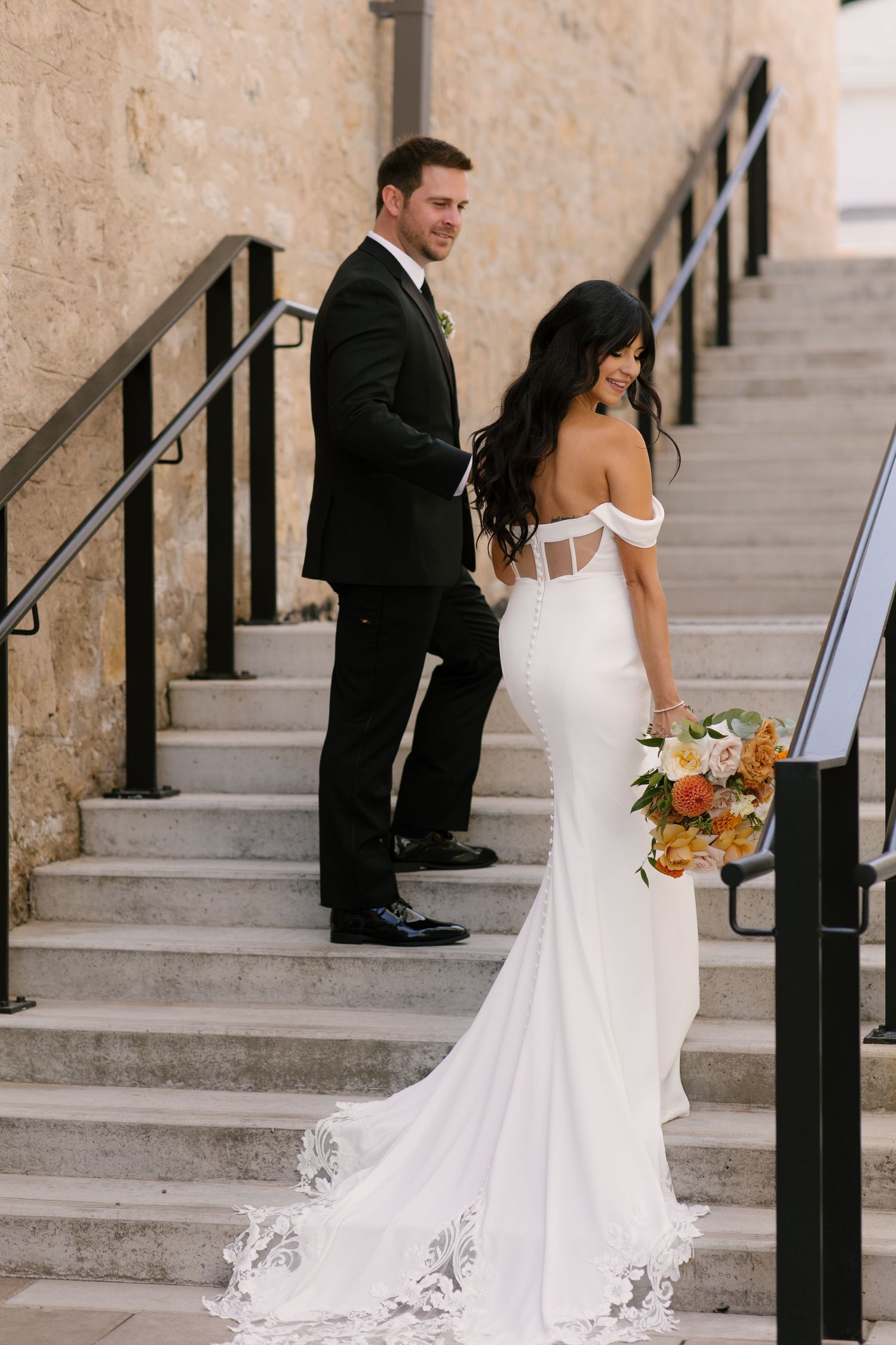 Bride in off-shoulder lace train gown and groom posing on stone outdoor staircase together.