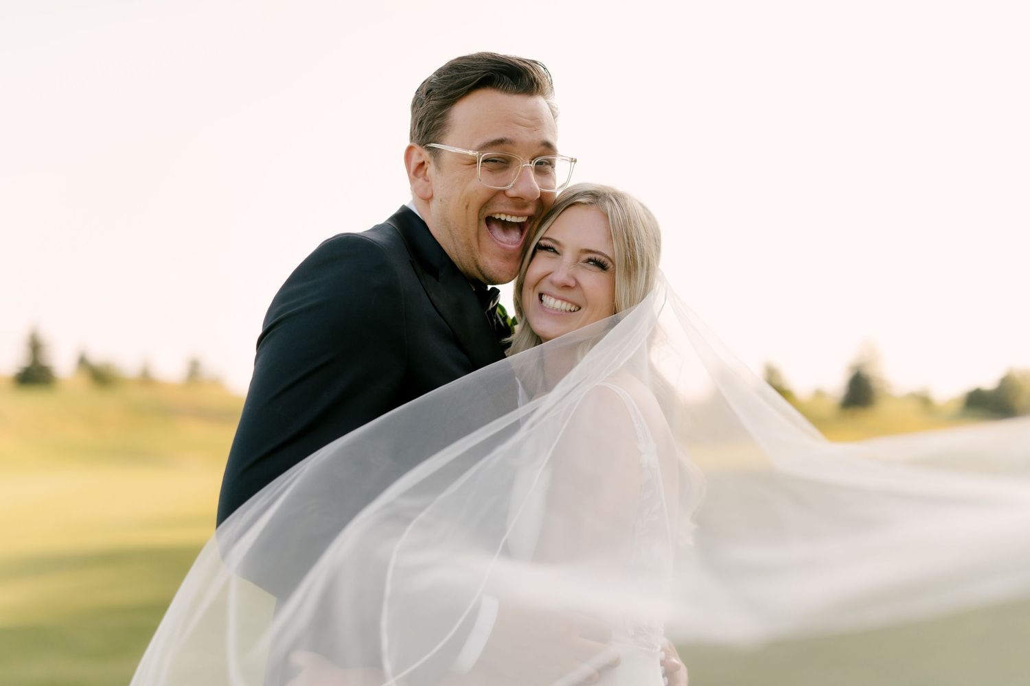 Joyful bride and groom laughing together as her veil blows in the wind at golden hour.