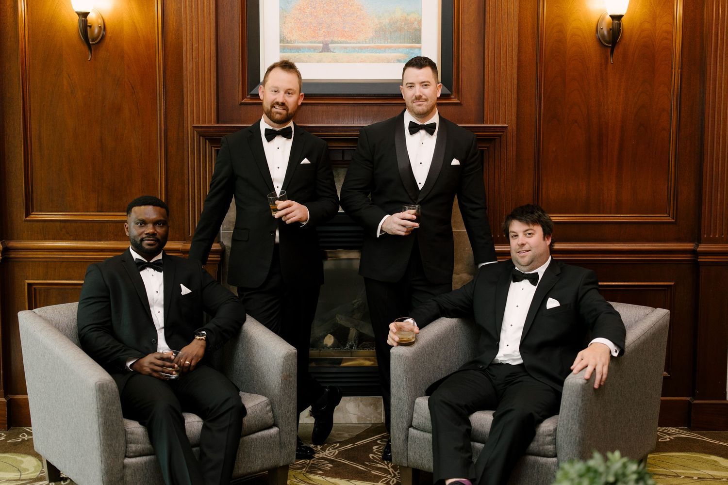Four groomsmen in black tuxedos relaxing in elegant wood-paneled room with grey lounge chairs.