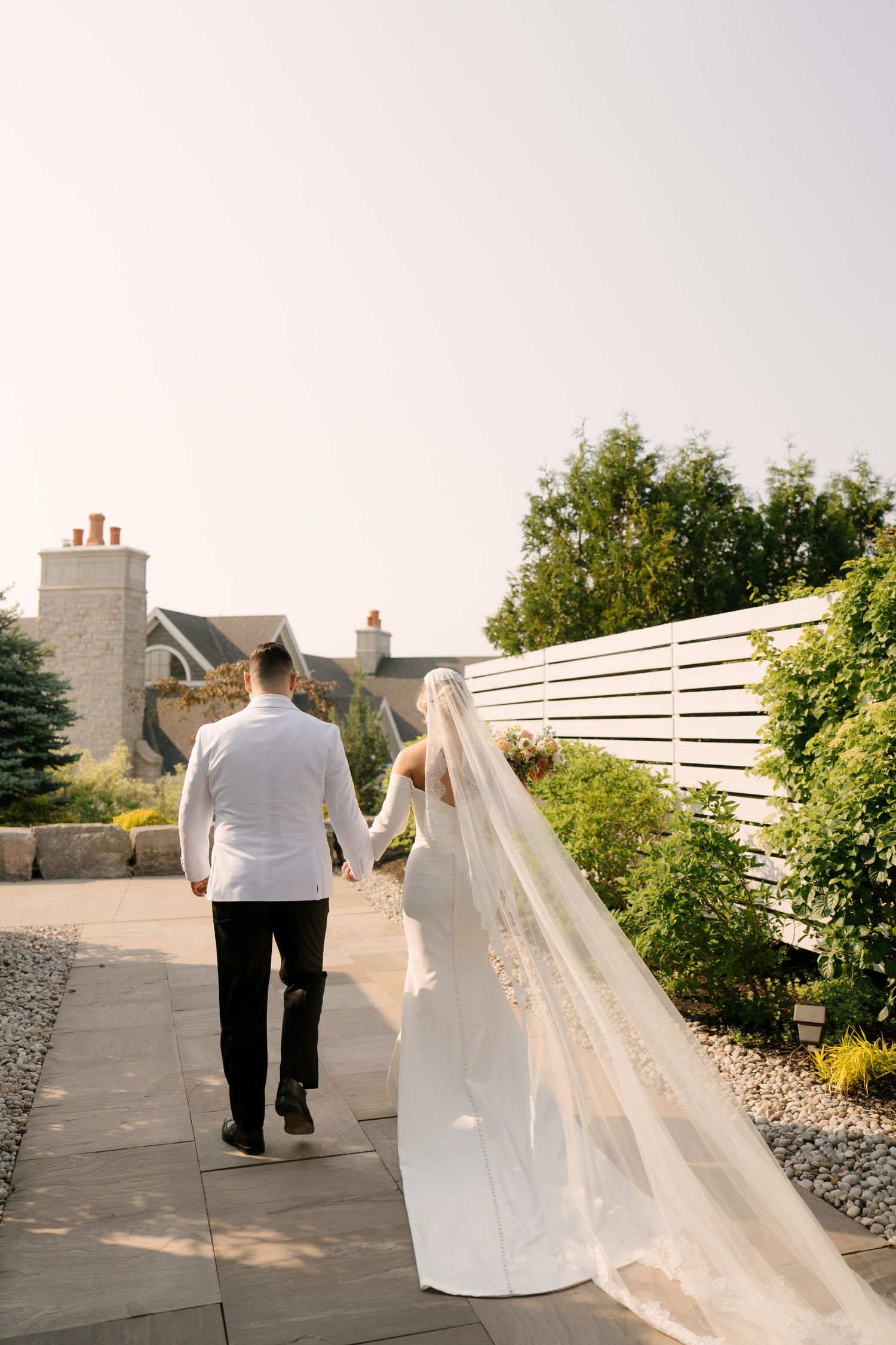 Bride and groom walking hand in hand on stone path, bride wearing long lace-trimmed cathedral veil trailing behind at Whistle Bear venue.