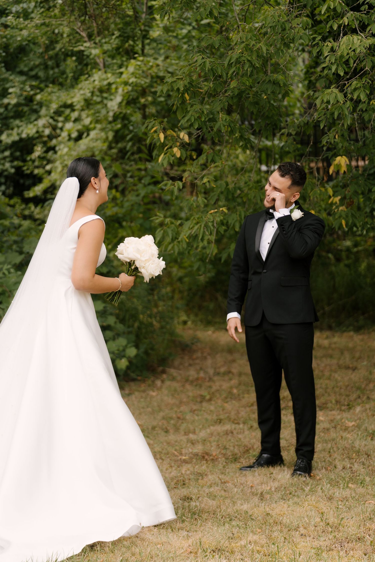 Groom wiping tears during emotional first look with bride holding white peony bouquet outdoors.