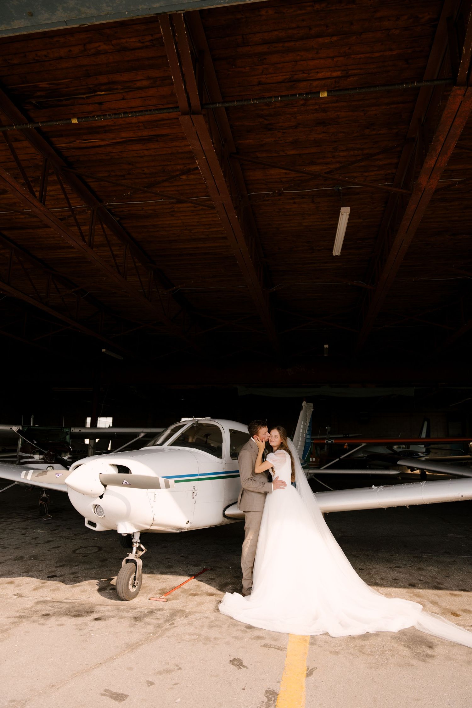 Bride and groom embrace in front of a small white aircraft inside a dimly lit airplane hangar.