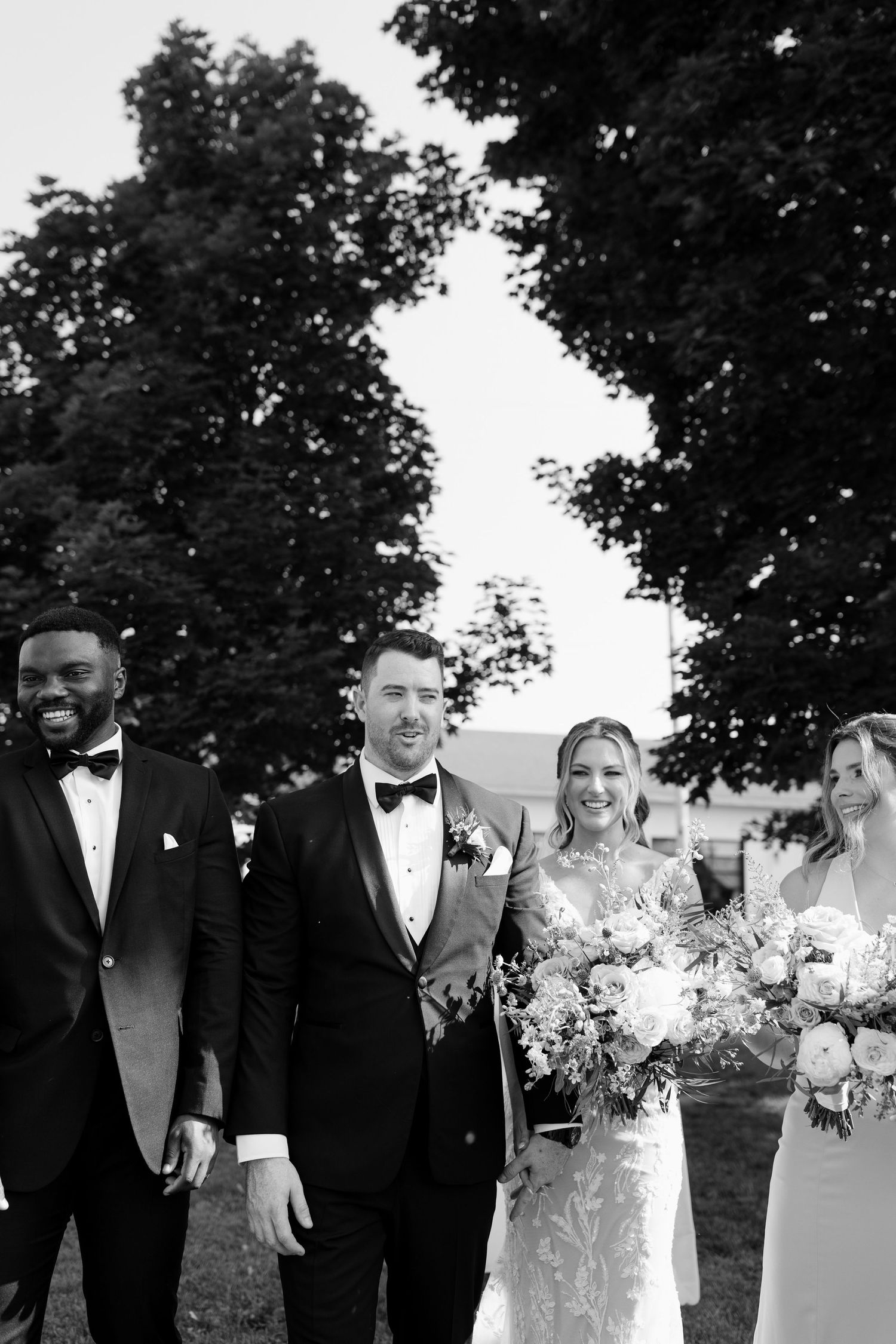 Black and white photo of smiling bride, groom, and wedding party walking together outdoors at St Marys venue.
