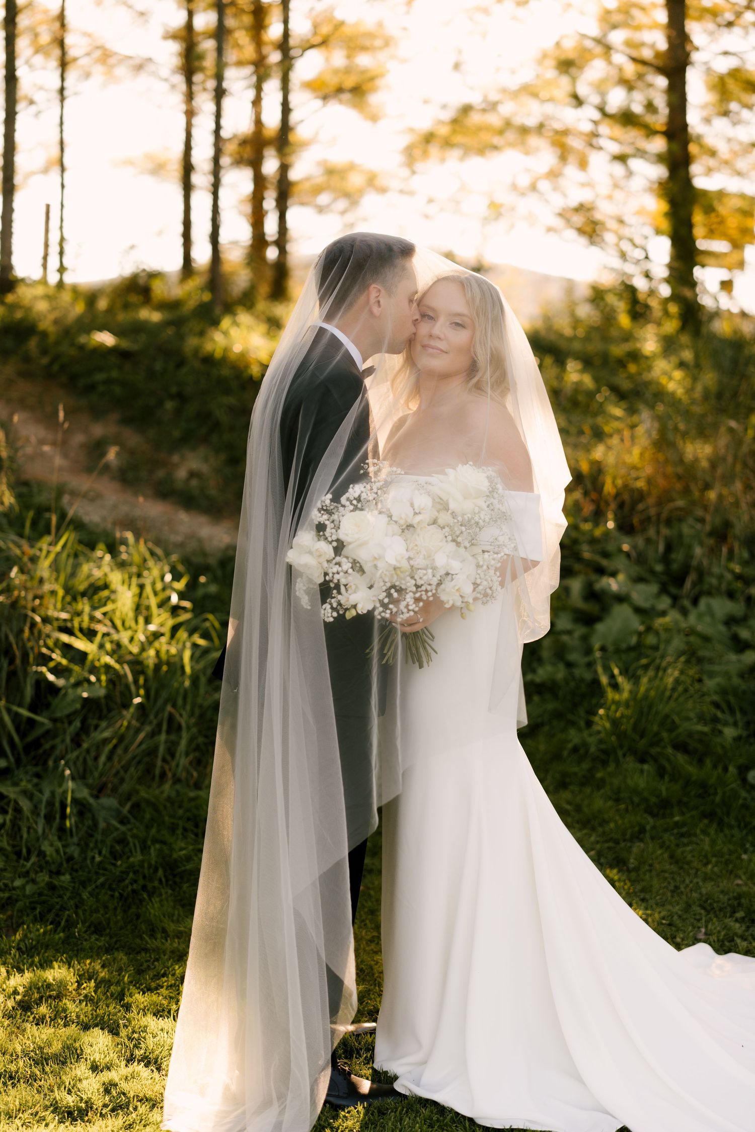Groom kisses bride on the cheek as she smiles holding a white floral bouquet in warm sunset light.