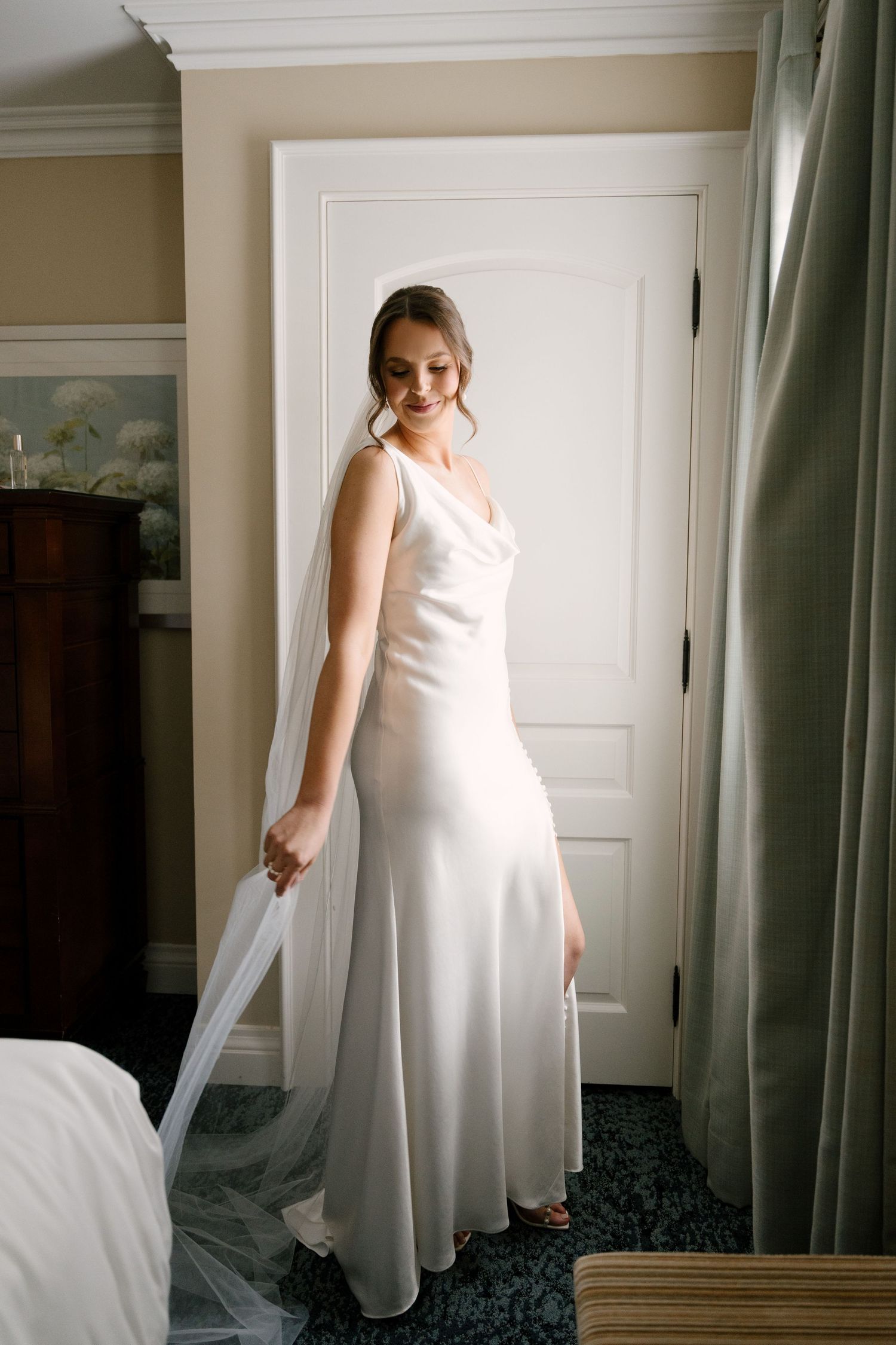 Smiling bride in sleek white satin gown holding veil in warmly lit hotel room.