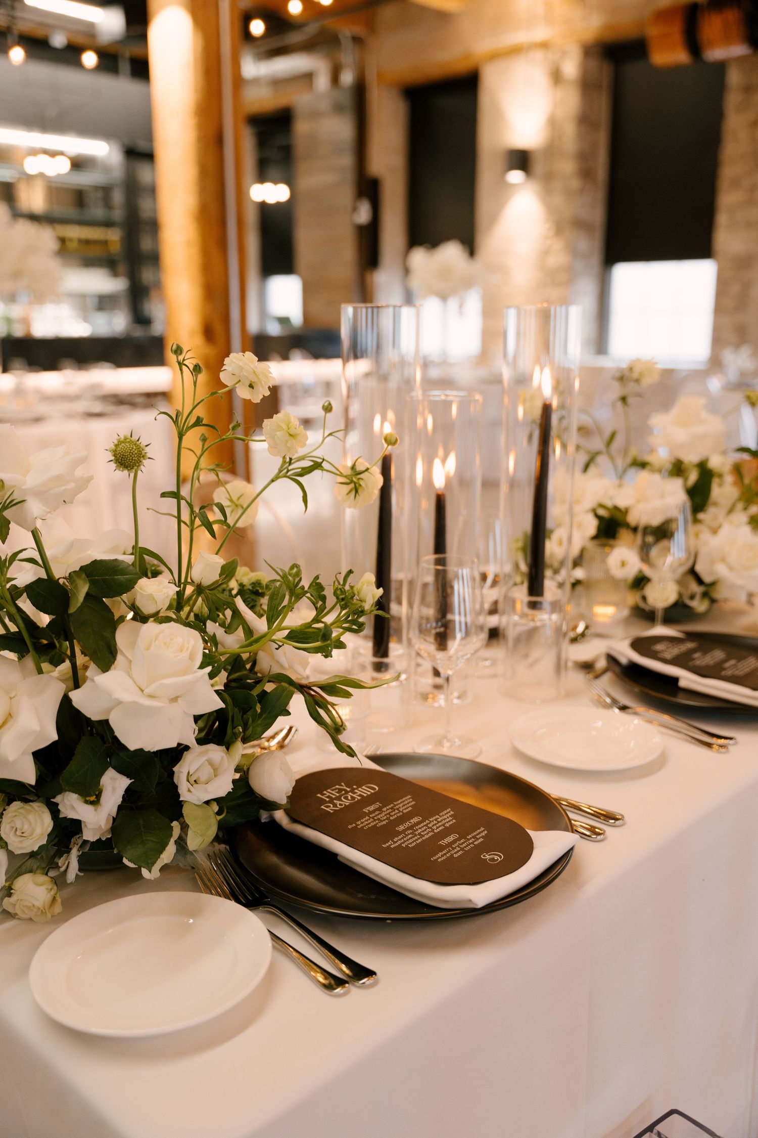 Close-up of wedding table setting with white roses, black taper candles, dark plates, gold cutlery, and a wooden menu card.