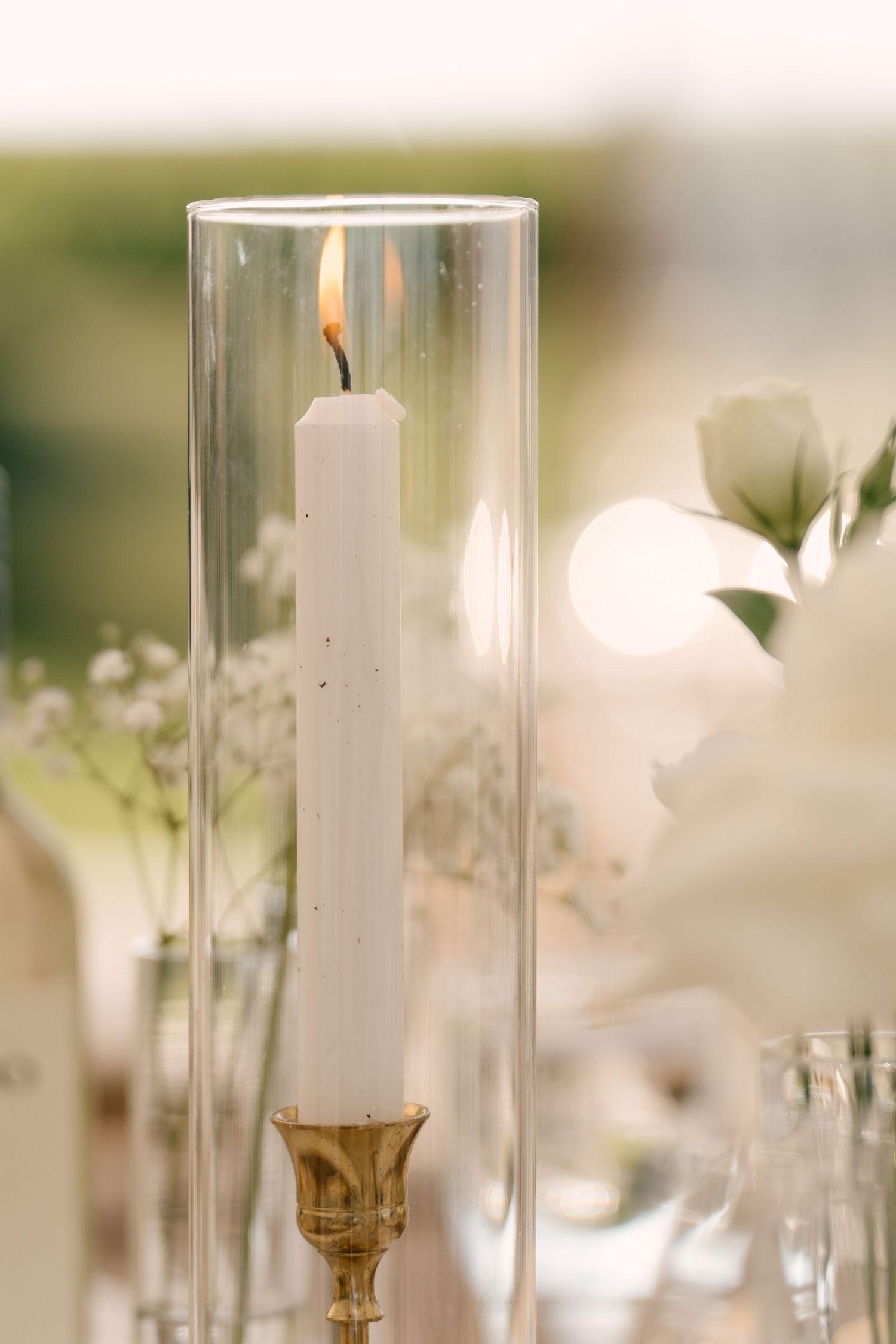 Lit white taper candle in glass hurricane holder on gold stand at elegant outdoor event table.