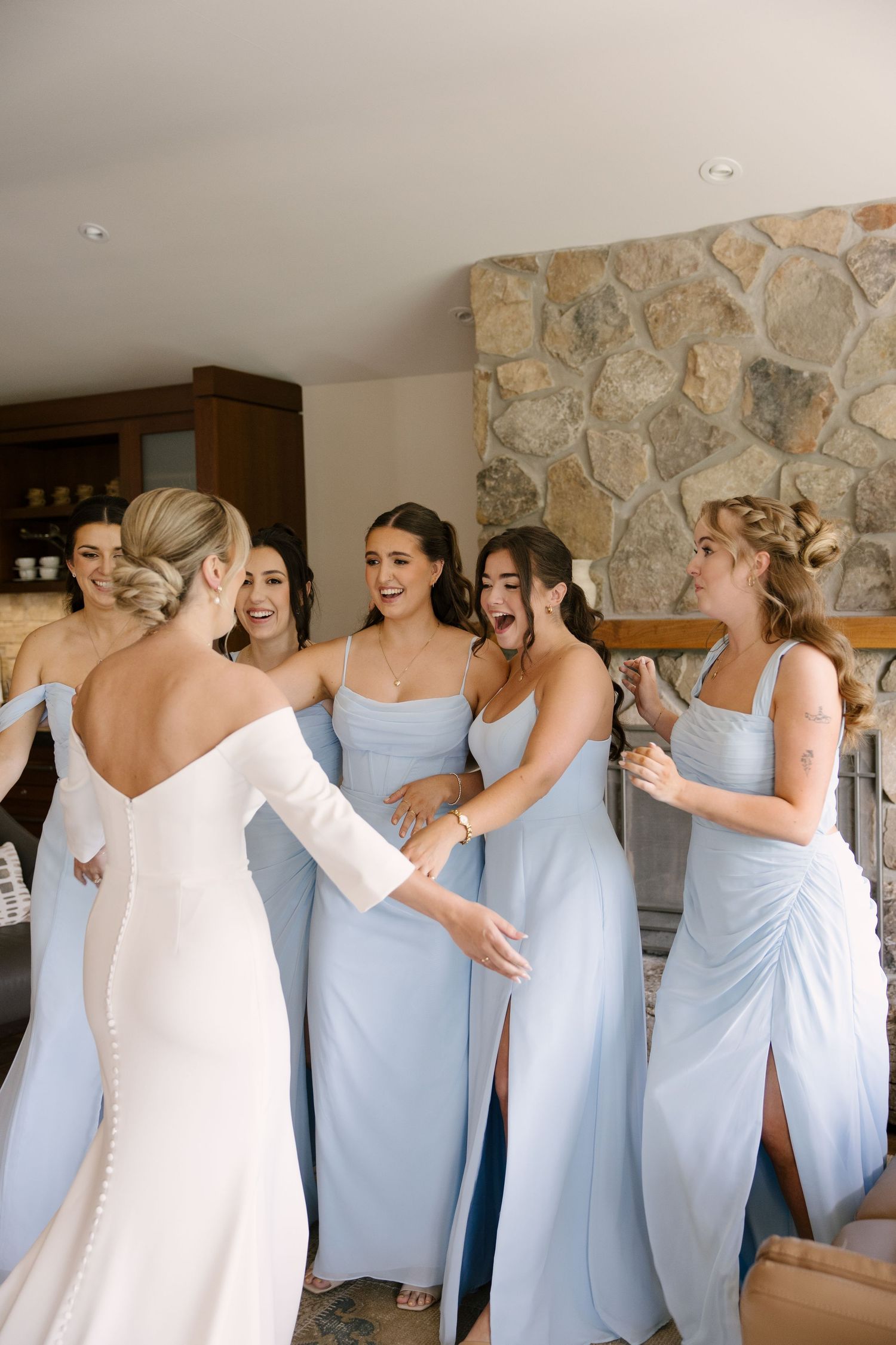 Bride in white off-shoulder gown greeted by excited bridesmaids in light blue dresses near a stone fireplace.