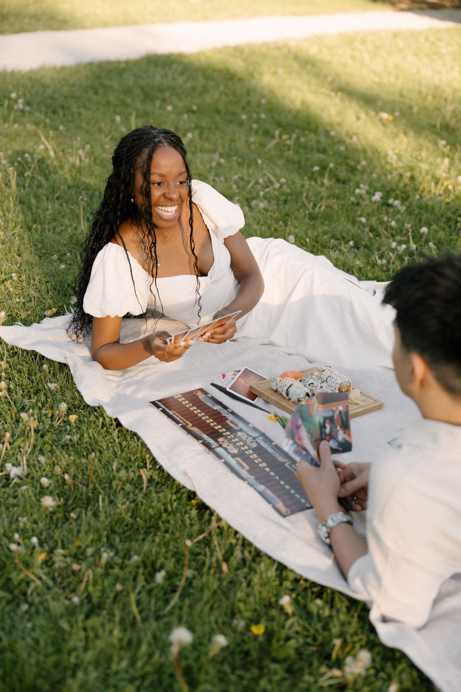 Smiling woman in white dress lying on blanket in park looking at man holding photos.
