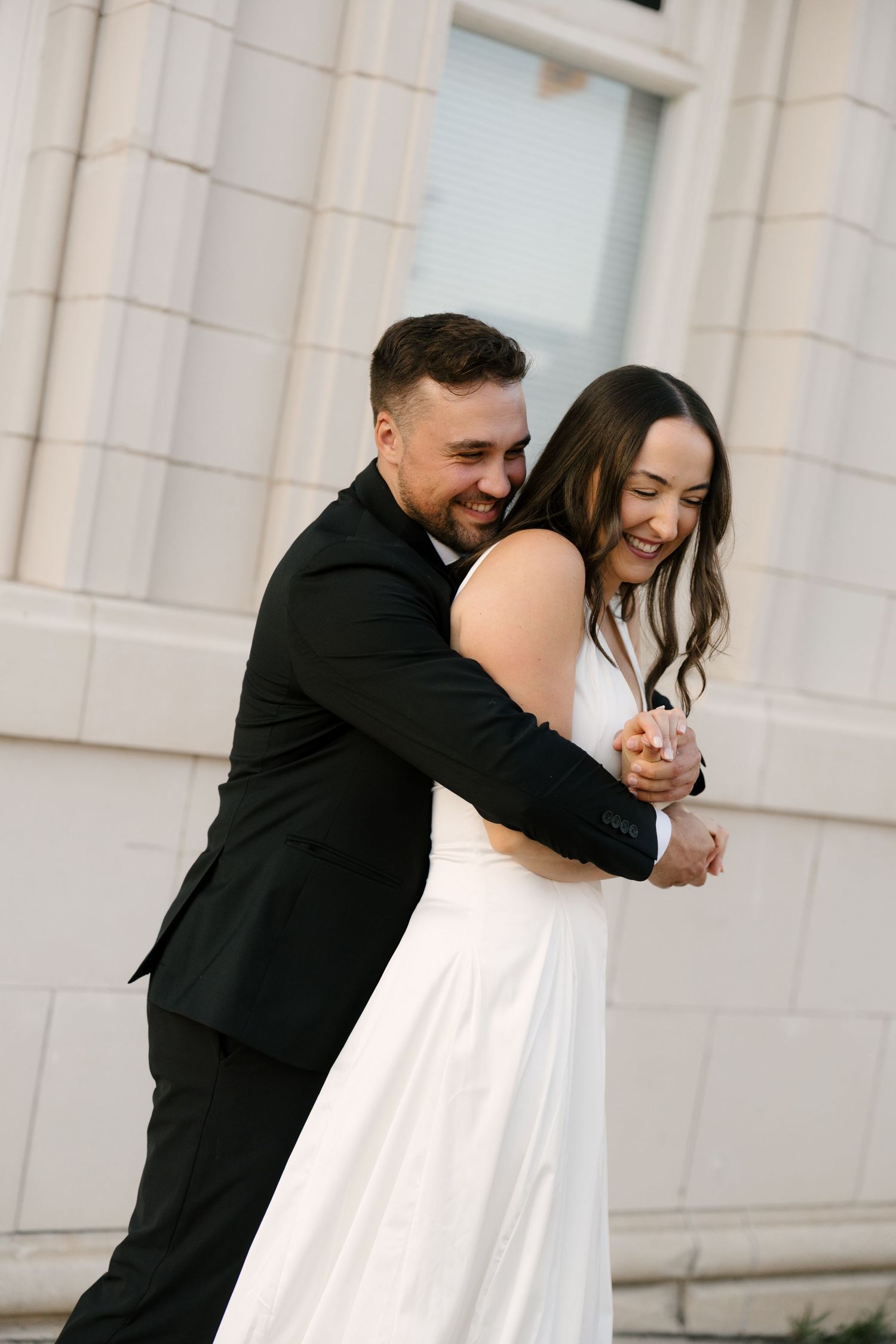 Groom in black suit embracing laughing bride in white dress during outdoor wedding photos.