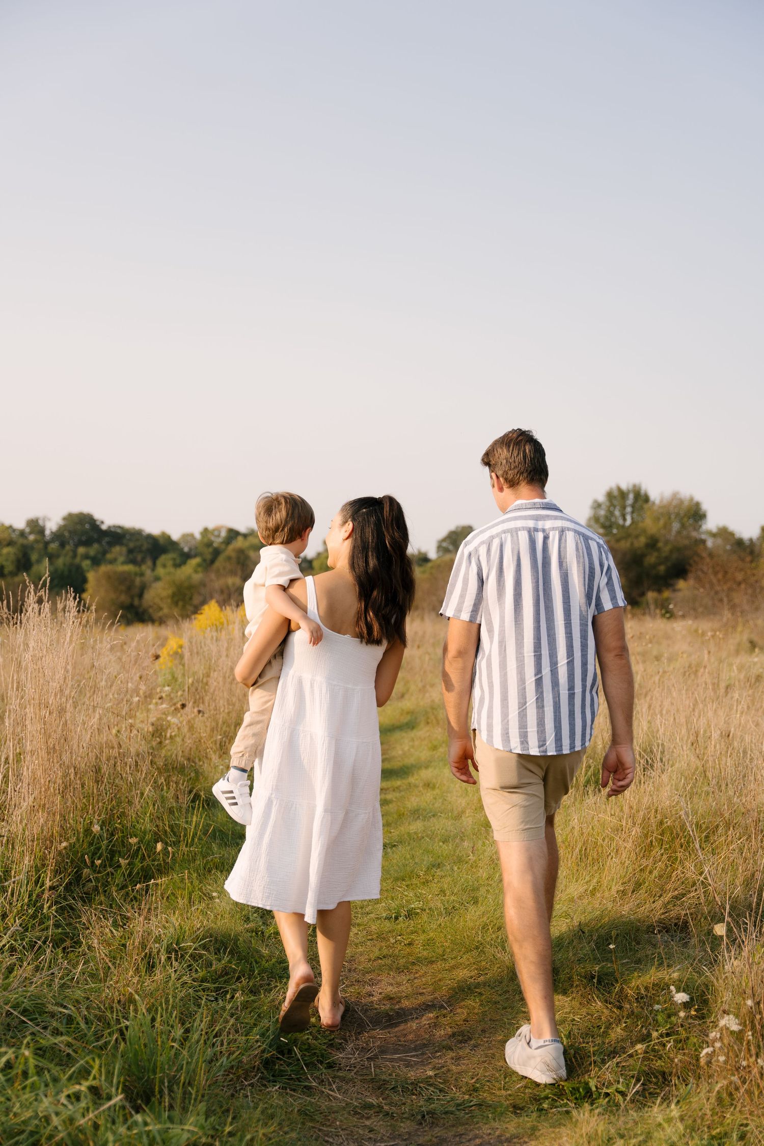 Family of three walks away along a meadow path at sunset, mother carrying toddler beside father.