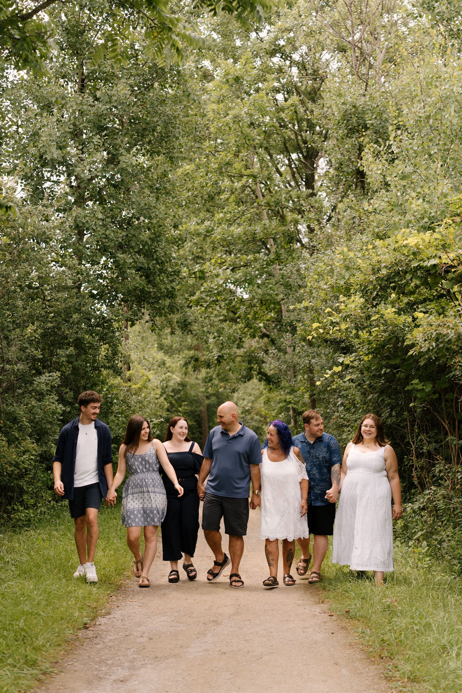 Extended family of seven walking hand-in-hand along a tree-lined path outdoors.
