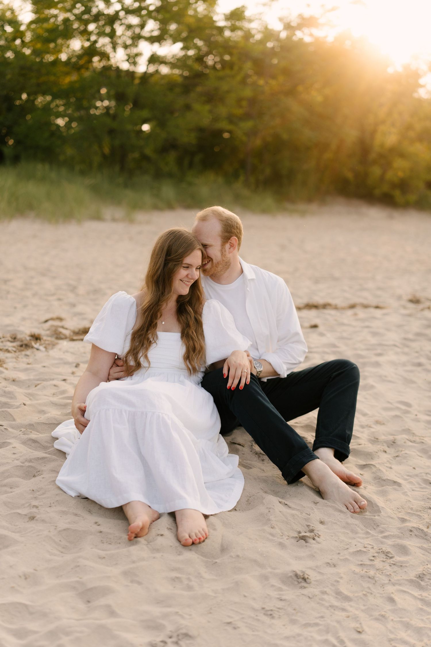 Couple sitting barefoot on sandy beach at sunset, leaning close together in coordinating white outfits.