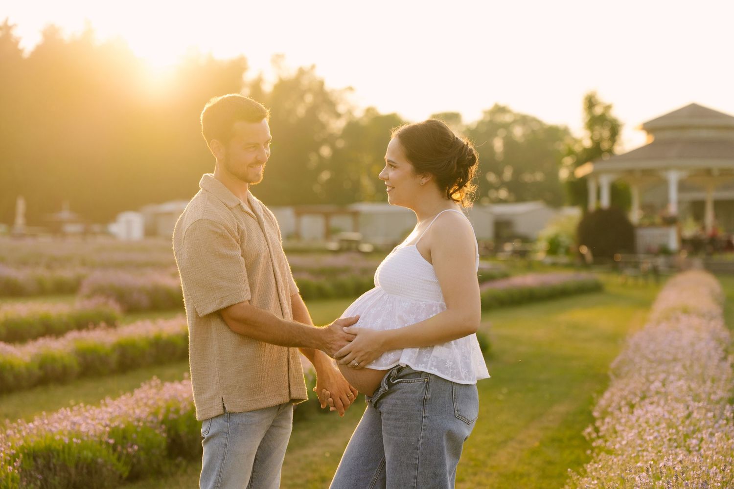 Couple holding hands in a lavender field at golden hour, pregnant woman in white top and jeans.