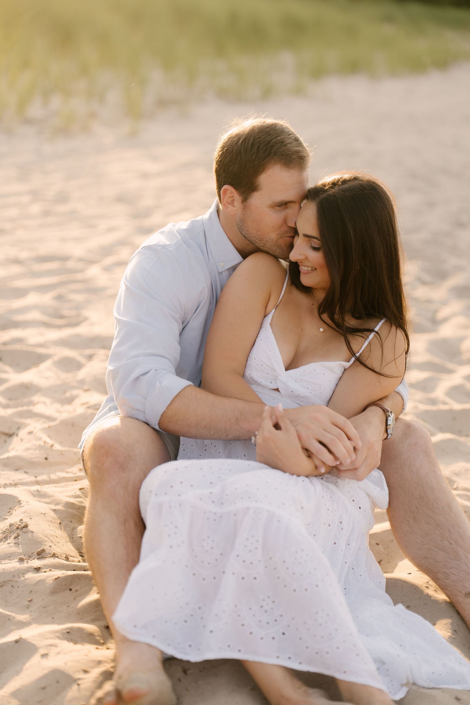 Romantic couple embracing on sandy beach at golden sunset, woman in white dress sitting in mans arms.