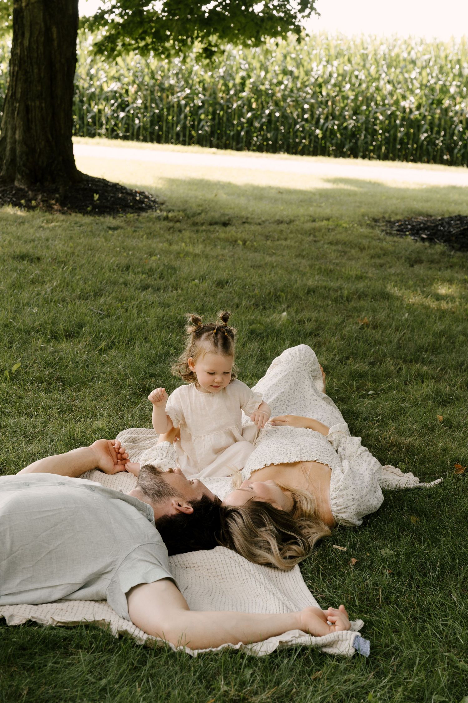 Two parents lie on a blanket in the grass while their toddler daughter sits between them outdoors.