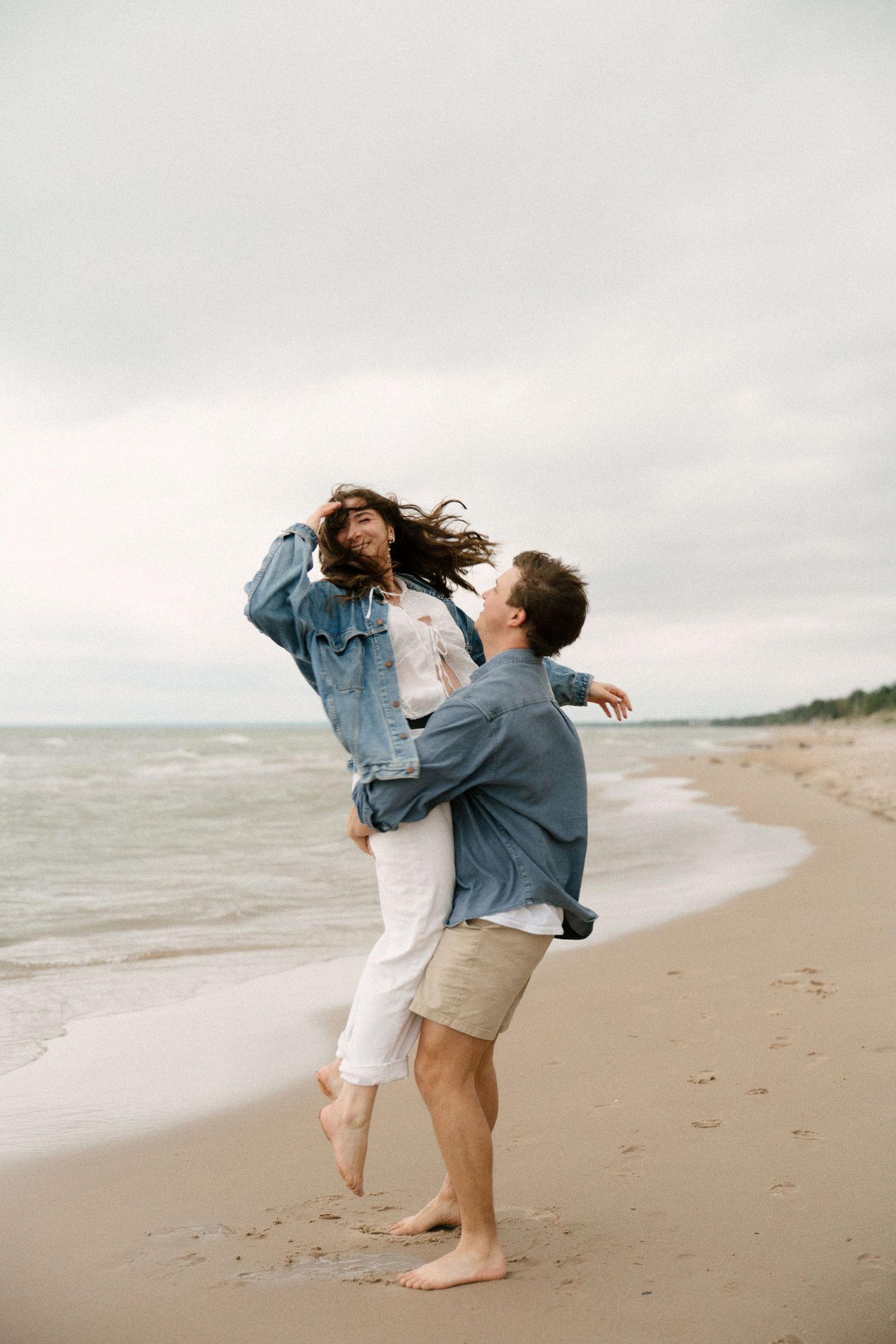 Couple laughing and spinning together barefoot on a sandy beach with ocean waves in the background.