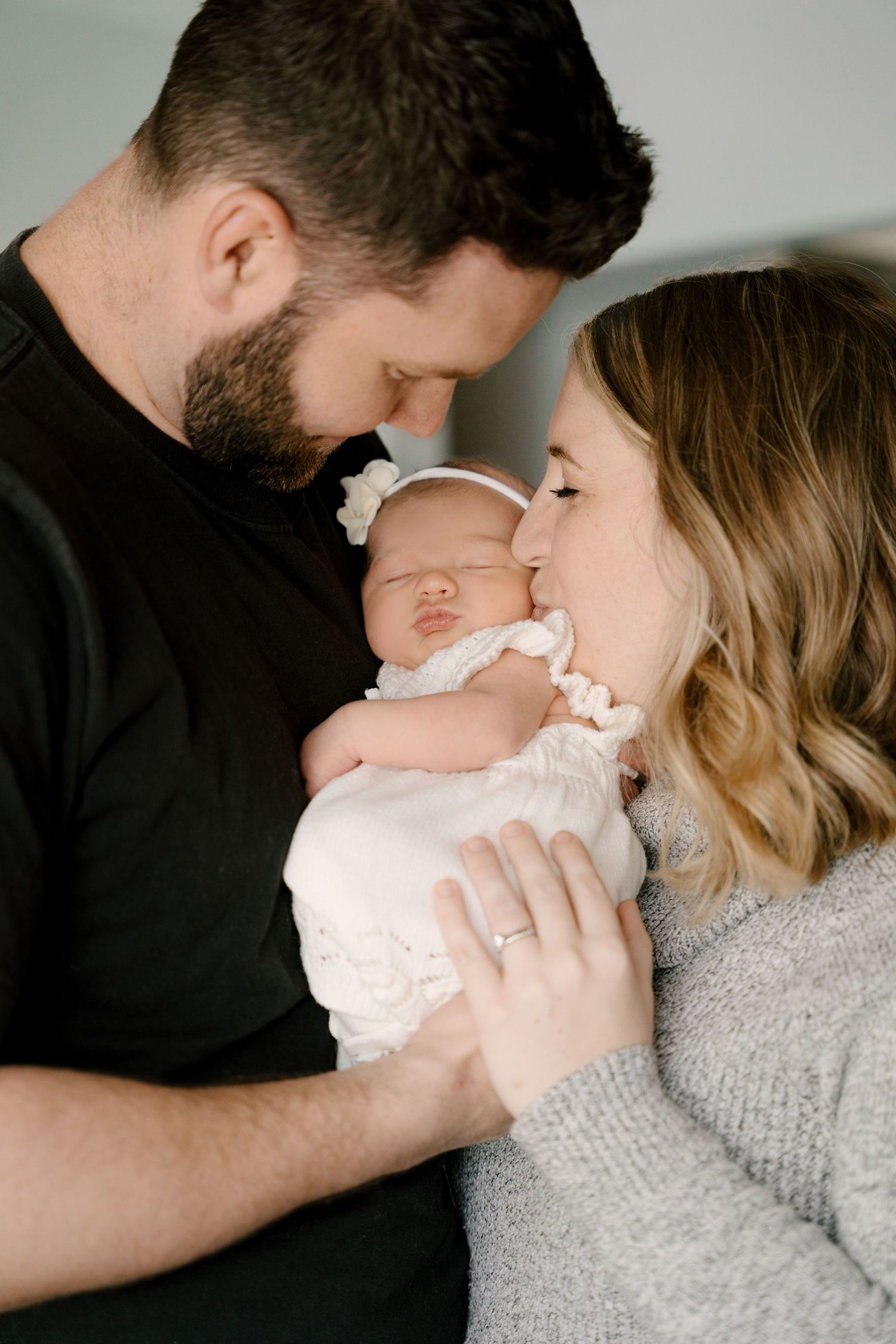 New parents tenderly touching foreheads over their sleeping newborn baby girl in white outfit.