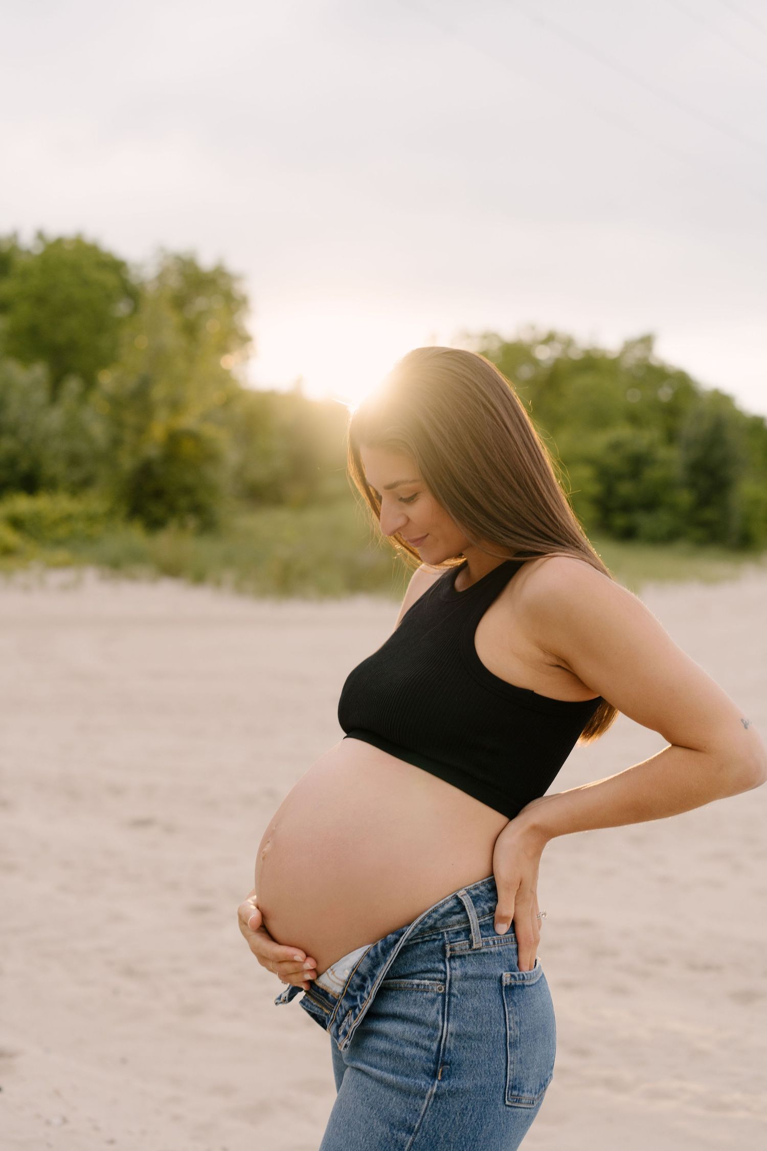 Pregnant woman in black crop top and jeans holding her baby bump at sunset on a sandy beach.