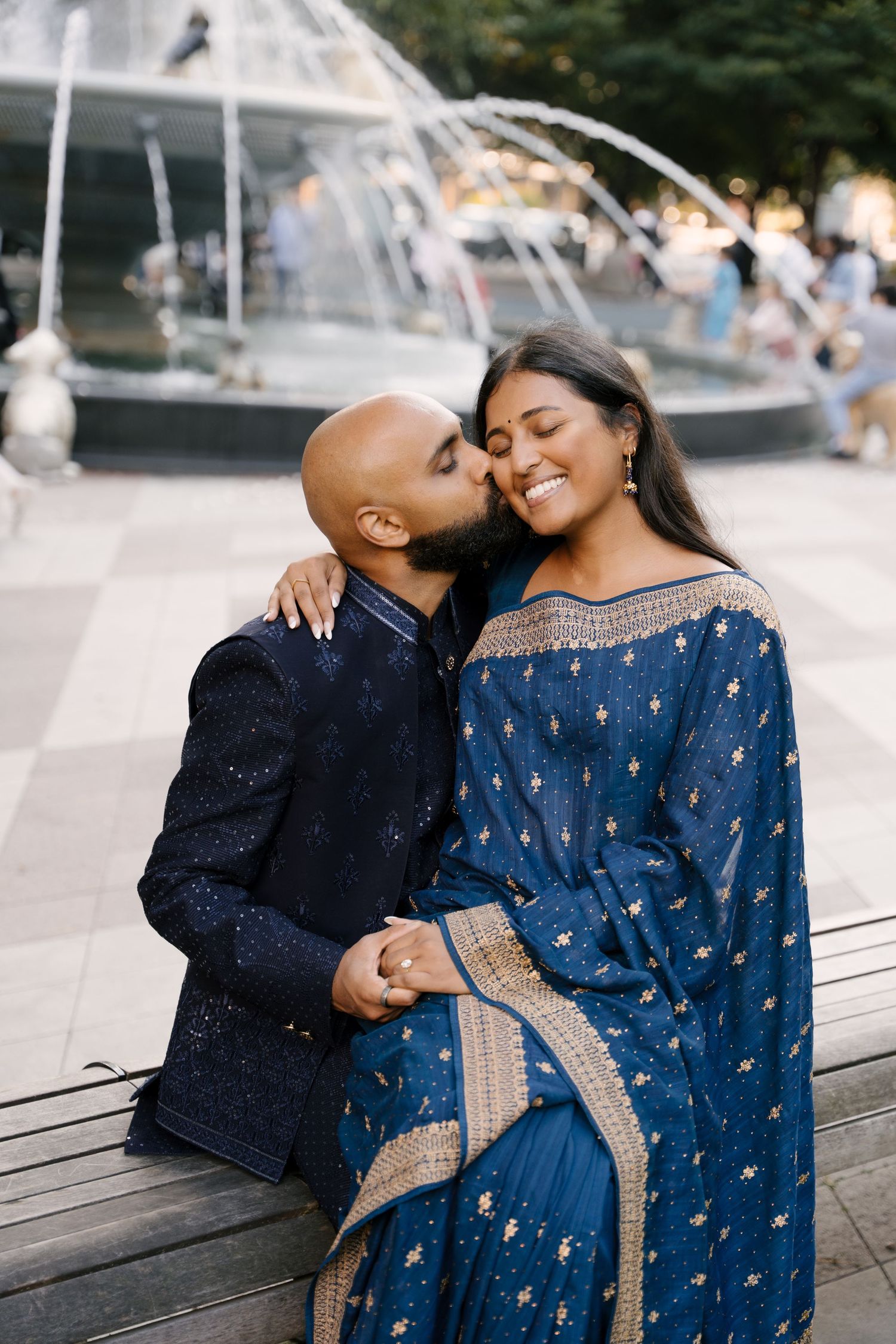 A couple in blue Indian attire shares a romantic kiss by a fountain, the woman smiling joyfully.