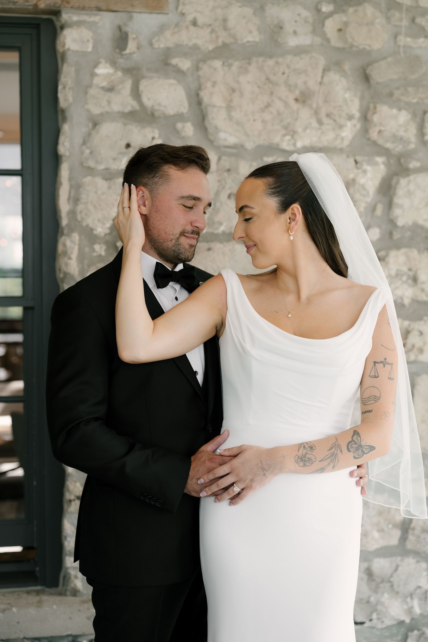 Bride and groom in elegant wedding attire share a romantic moment against a stone wall backdrop at Cambridge Mill.