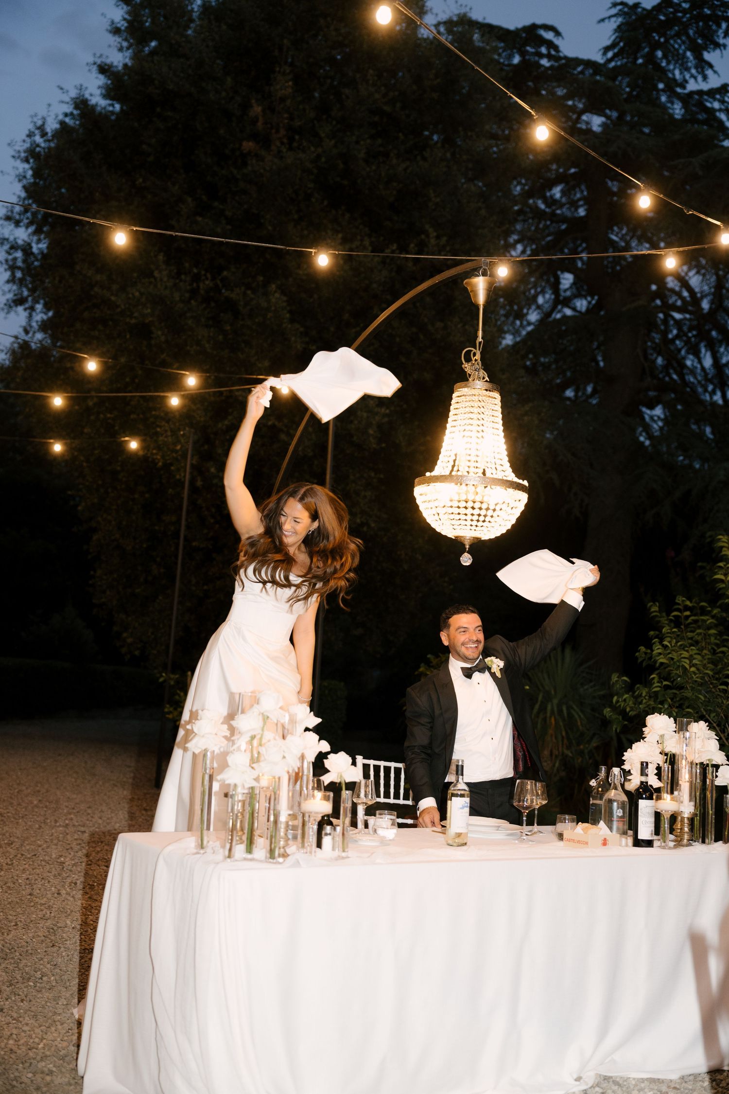 Bride and groom waving white napkins at outdoor wedding reception table under crystal chandelier.