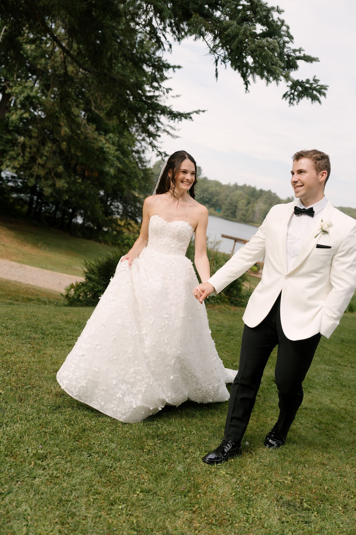 A bride in a strapless lace gown and groom in white tuxedo hold hands walking on green lawn near a lake.