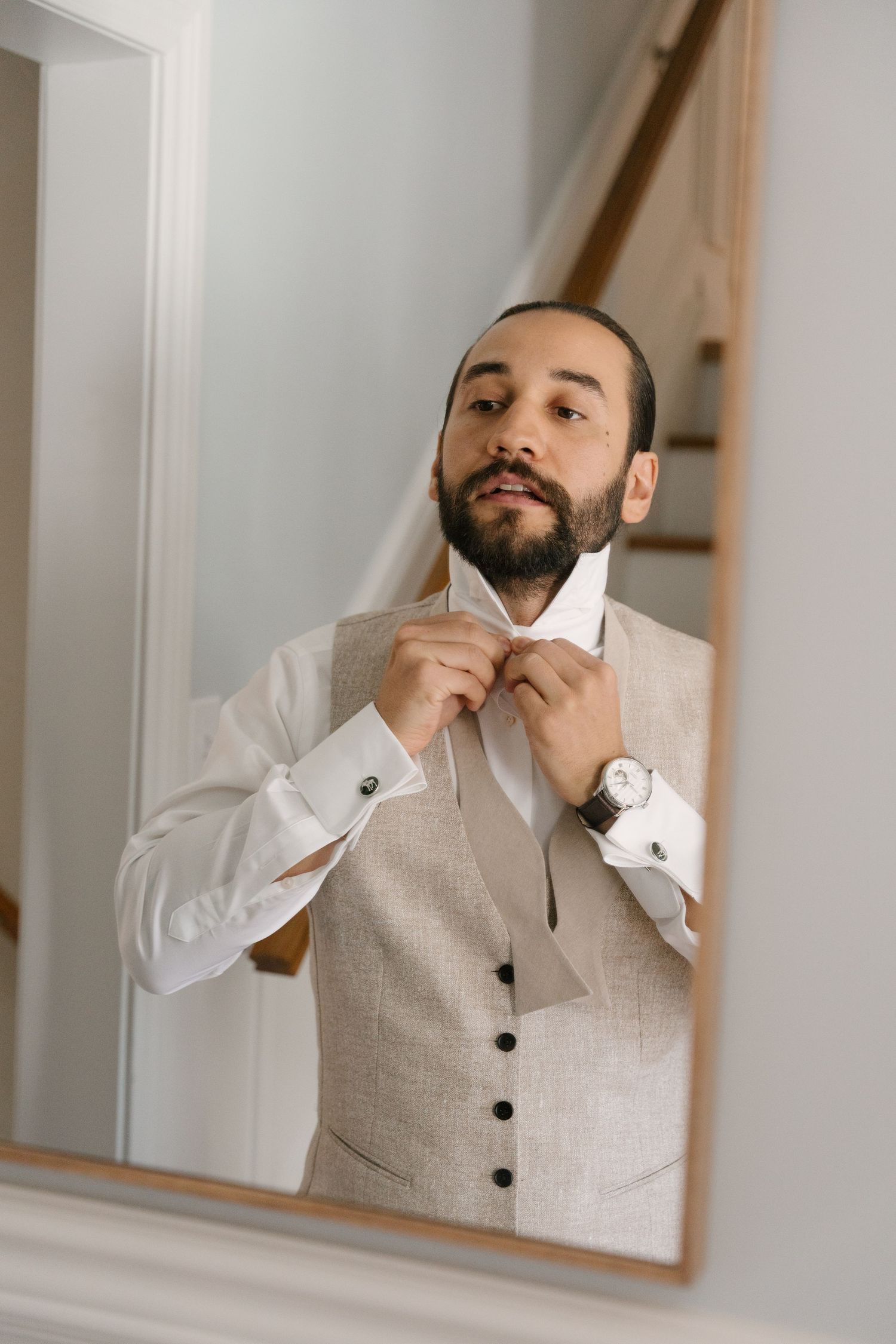 Bearded groom in beige vest adjusting white tie in mirror reflection before wedding ceremony.