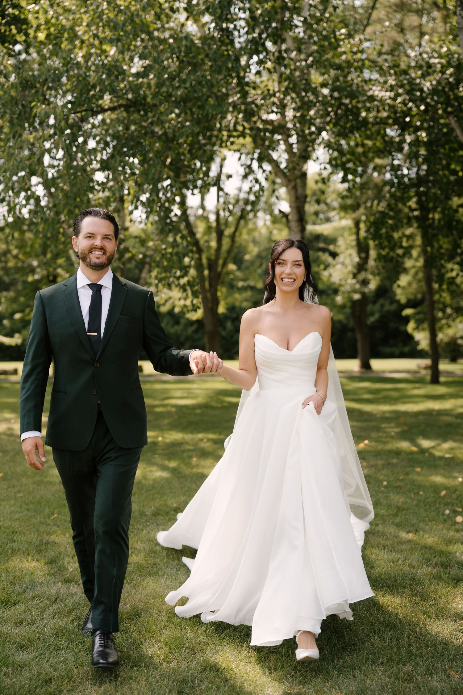 Smiling bride and groom in green suit walking hand in hand through lush garden at Roseville Estates wedding venue.