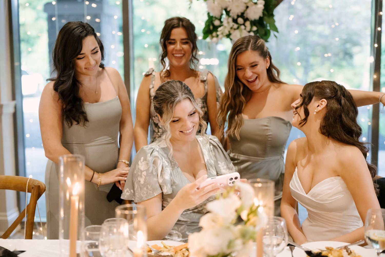 Bride smiles at phone surrounded by laughing bridesmaids in grey dresses at candlelit wedding reception table.
