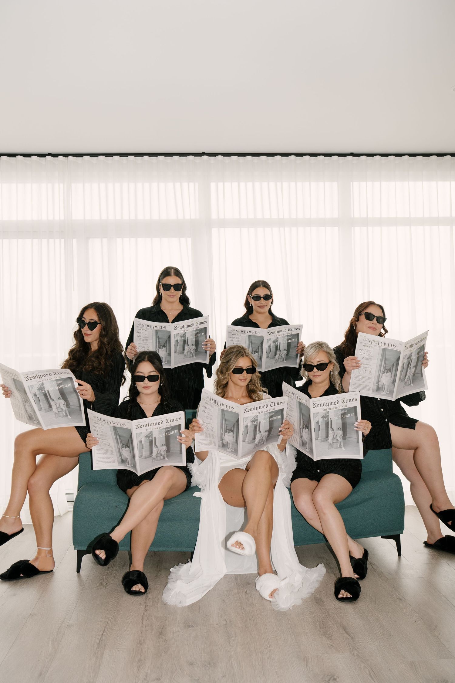 Bride and bridesmaids in black robes and sunglasses reading Newlywed Times newspapers while seated.