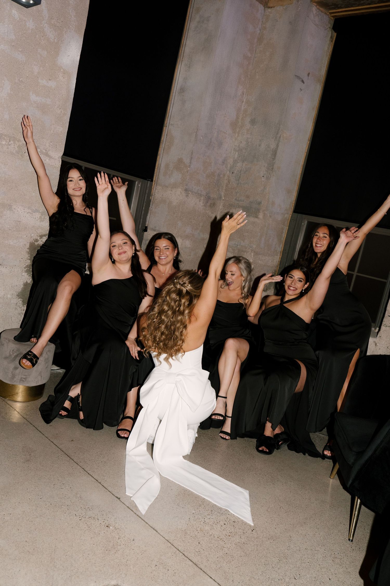 Bride in white bow-back dress tosses bouquet to excited bridesmaids in black gowns at wedding reception.