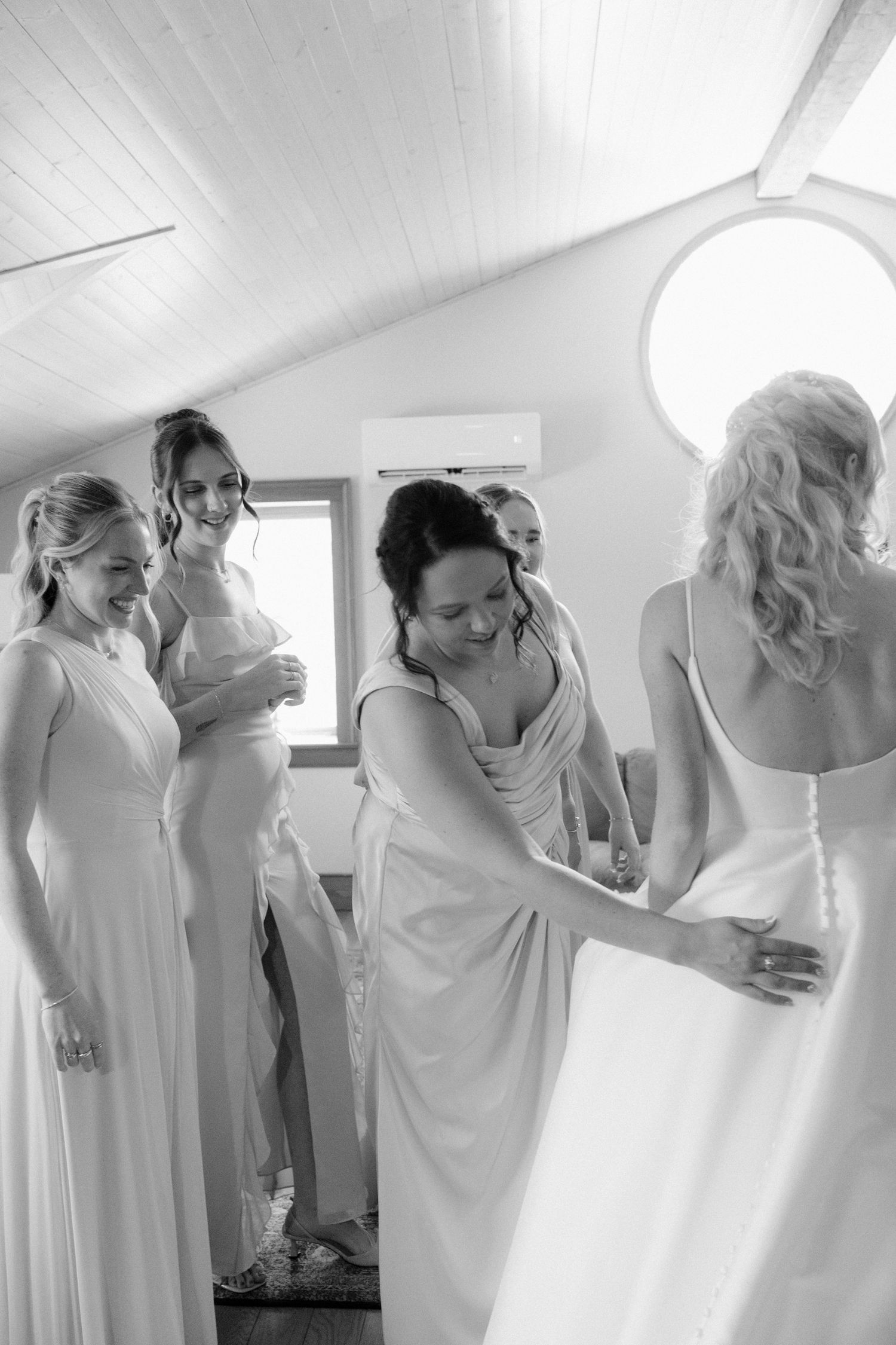 Bridesmaids help bride into her wedding gown in a bright white room before the ceremony.