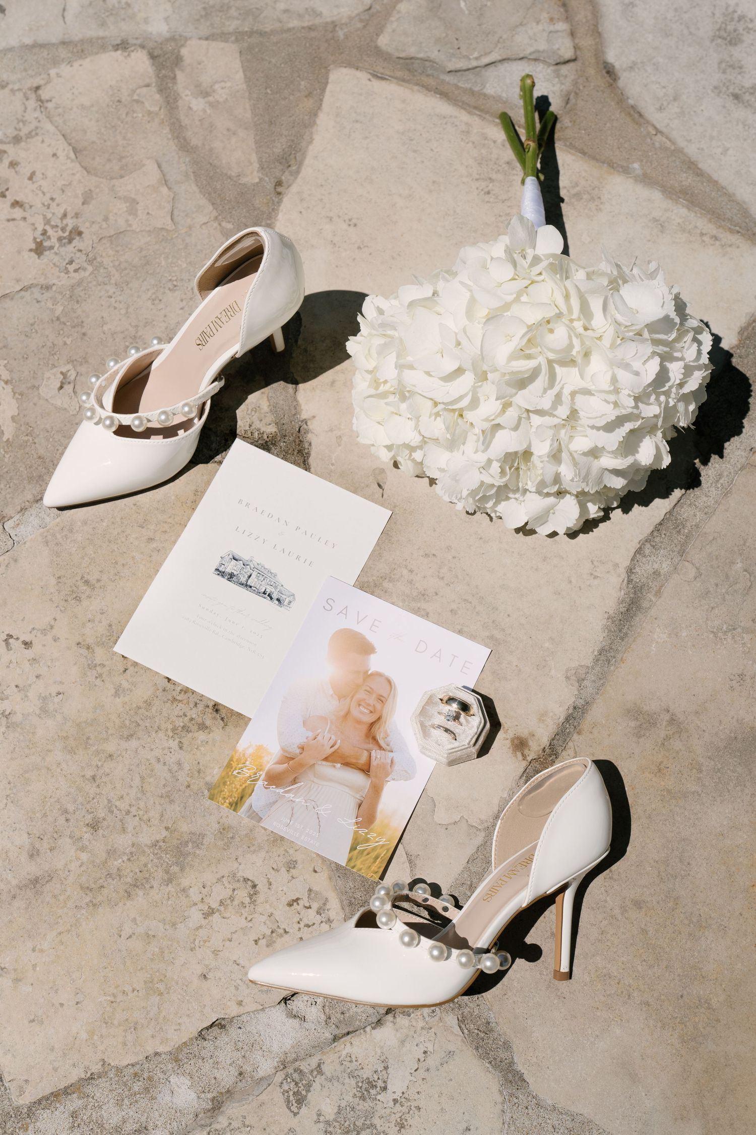 Flat lay of white bridal heels, wedding bouquet, save the date cards, and accessories on stone.