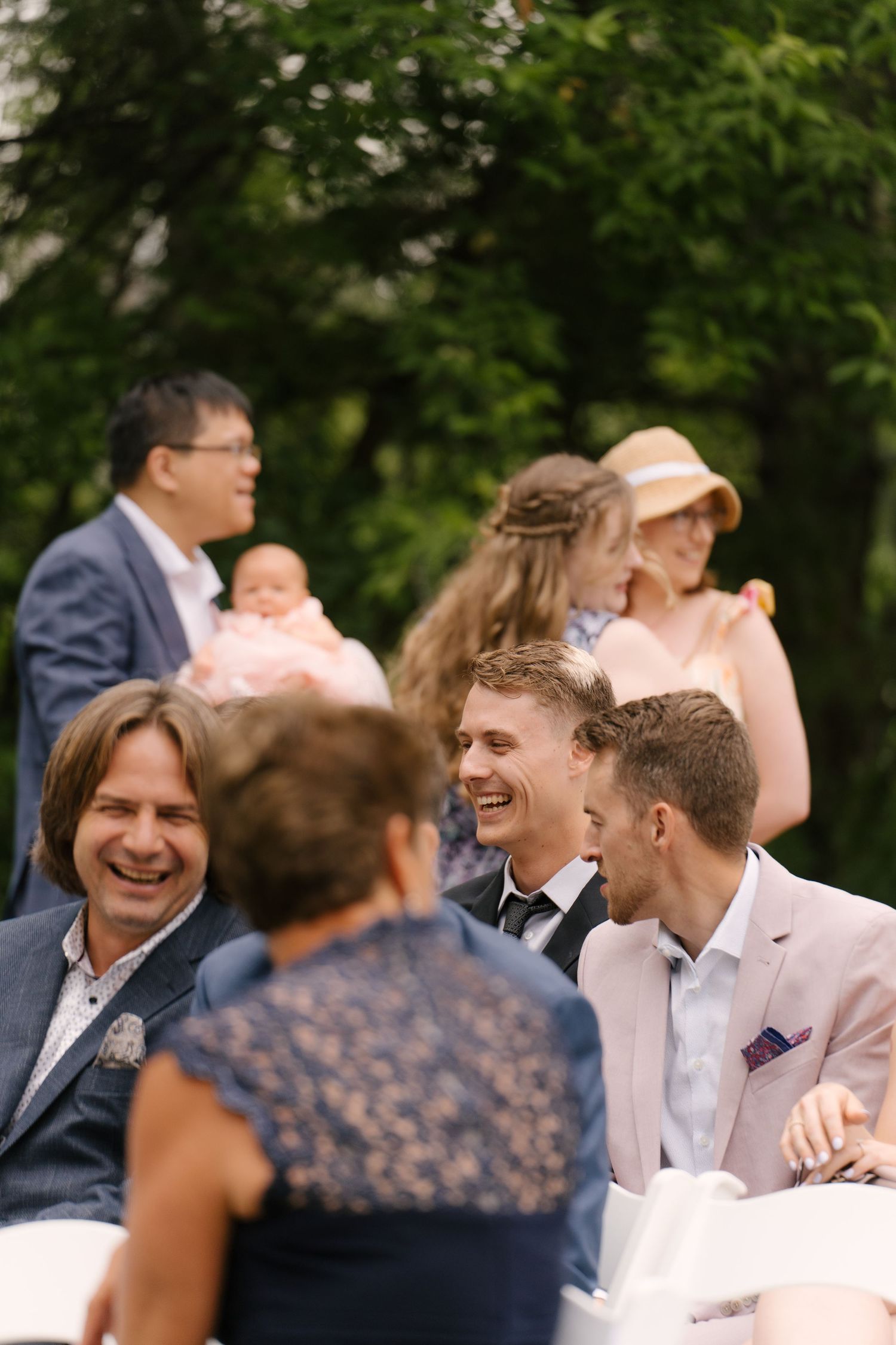 Wedding guests laugh and socialize joyfully while seated outdoors during a garden wedding ceremony.