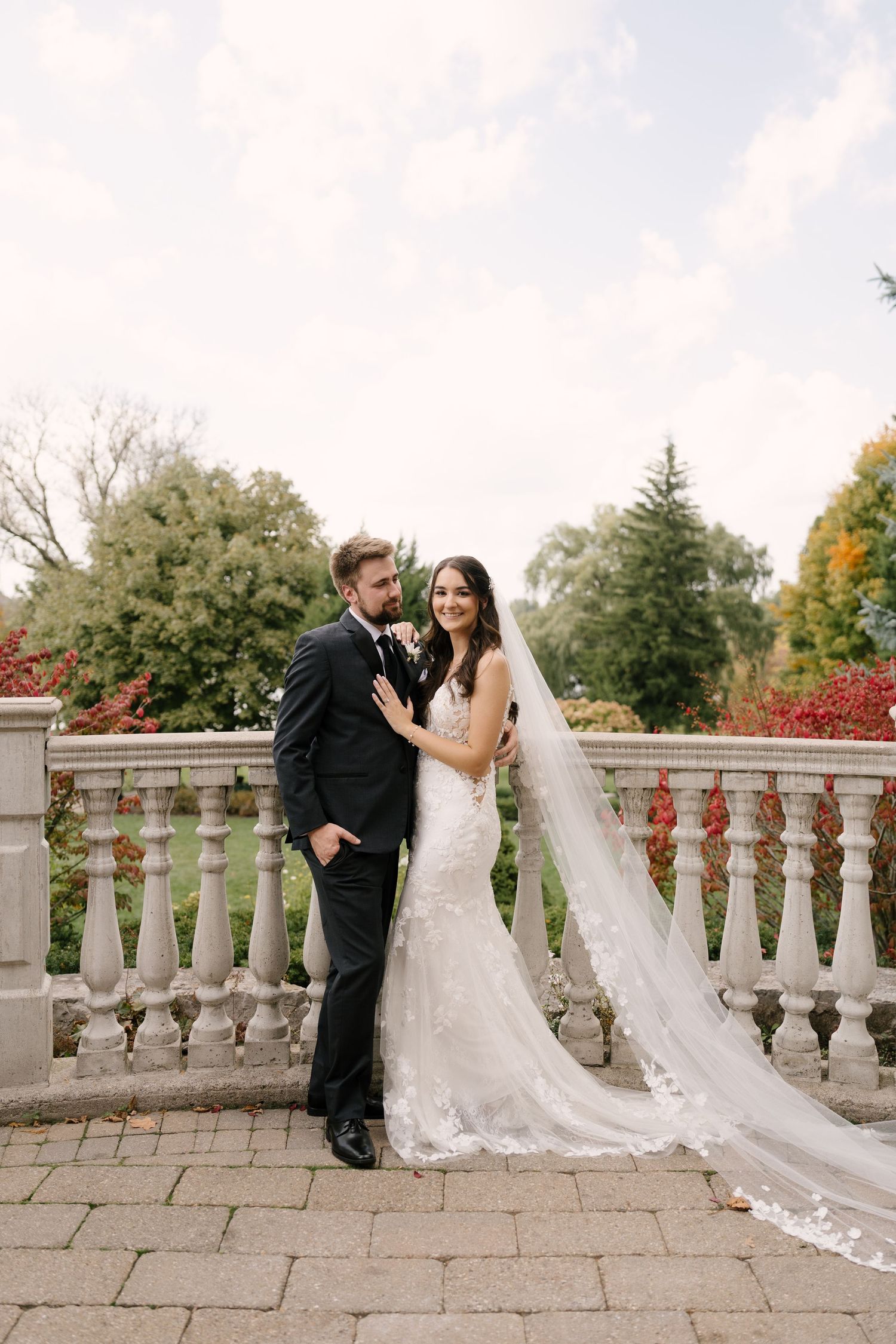 Bride and groom posing elegantly on a stone terrace with lace cathedral veil and autumn foliage backdrop.