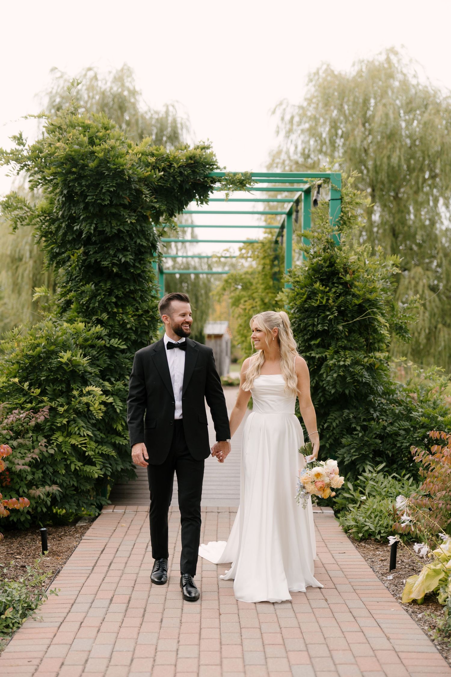 Bride and groom walking hand in hand through a lush garden pergola pathway on their wedding day at Pillar and Post.