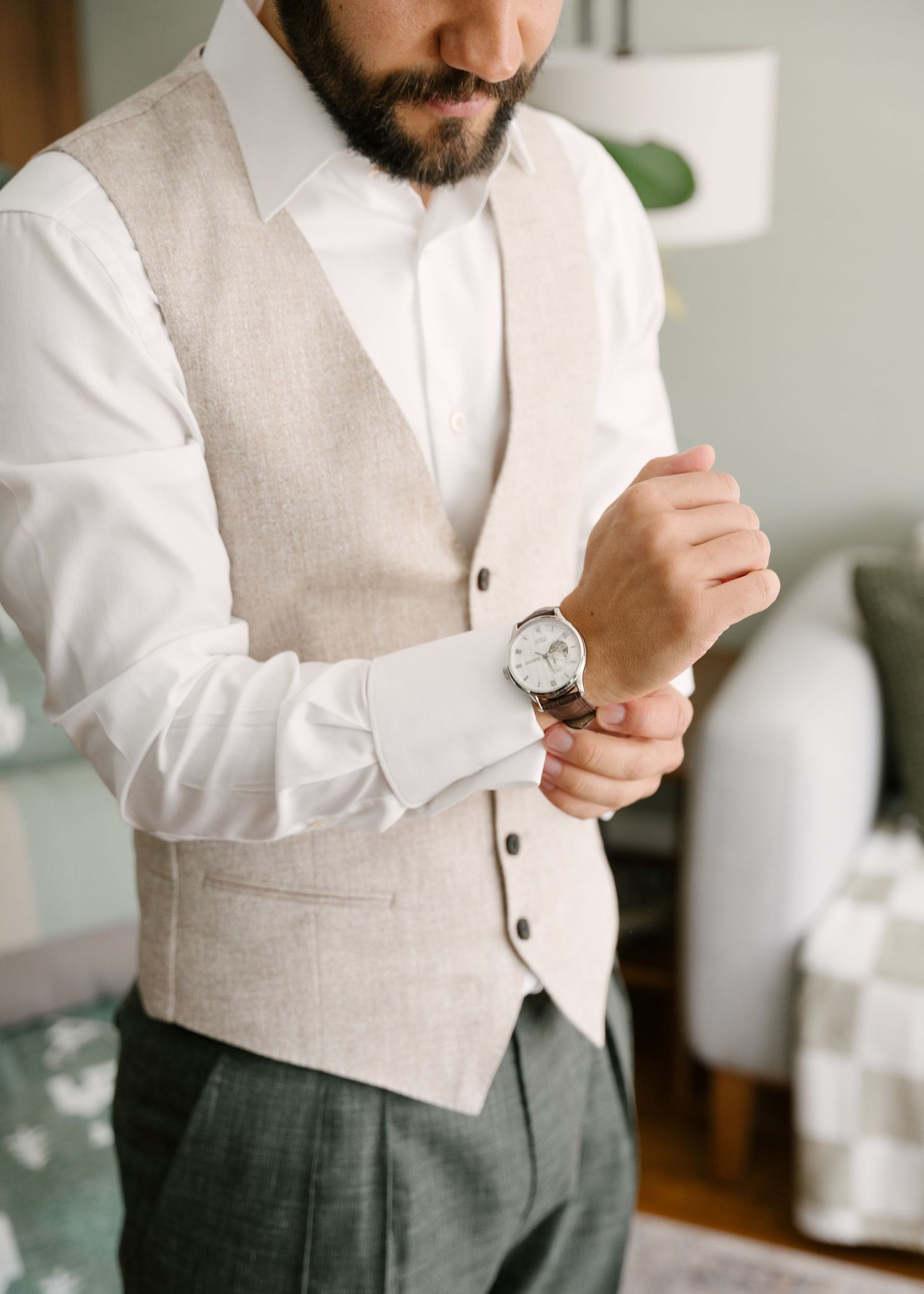 Bearded groom in beige vest and white dress shirt adjusting his classic silver watch while getting ready.