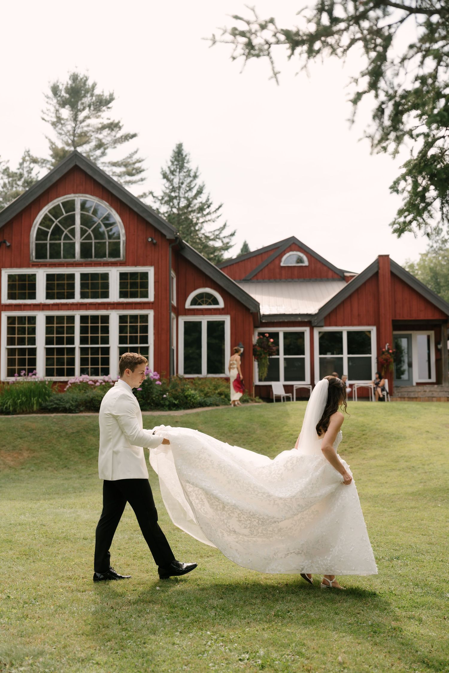 Groom in white jacket carries bride's lace wedding dress train as they walk across green lawn before red barn venue.