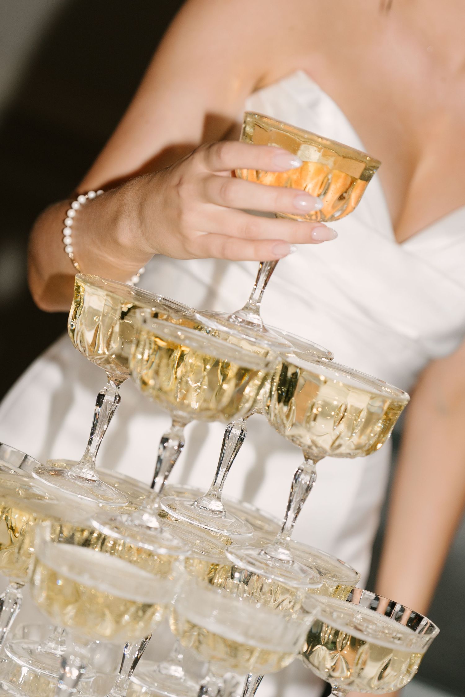 Bride lifting a champagne coupe from an elegant crystal tower of glasses at a wedding reception.