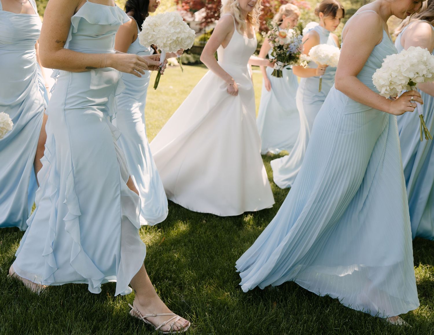 Bridesmaids in light blue dresses and bride in white gown walking across green grass at a wedding.