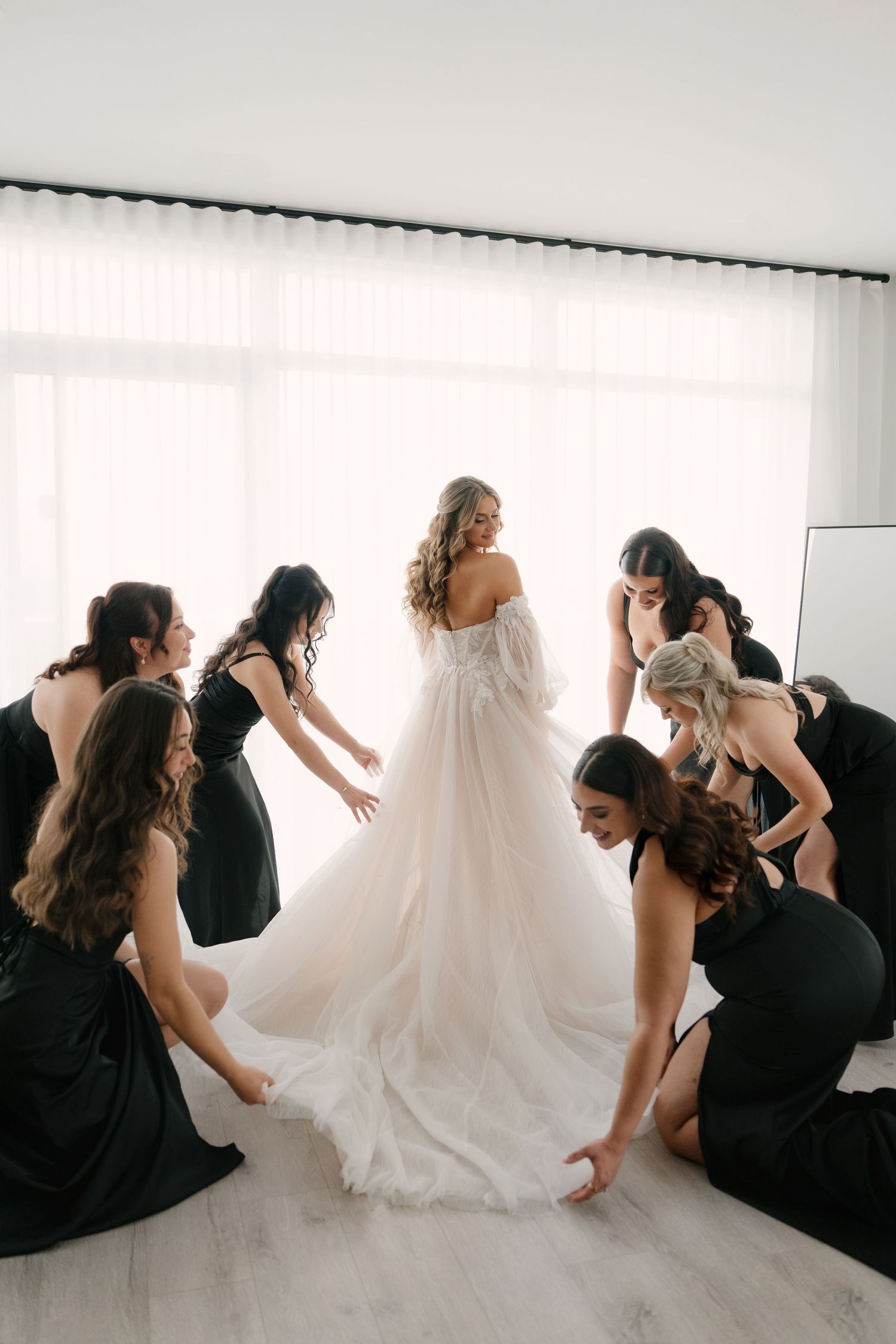 Bridesmaids in black dresses help bride into flowing white tulle wedding gown by bright window.