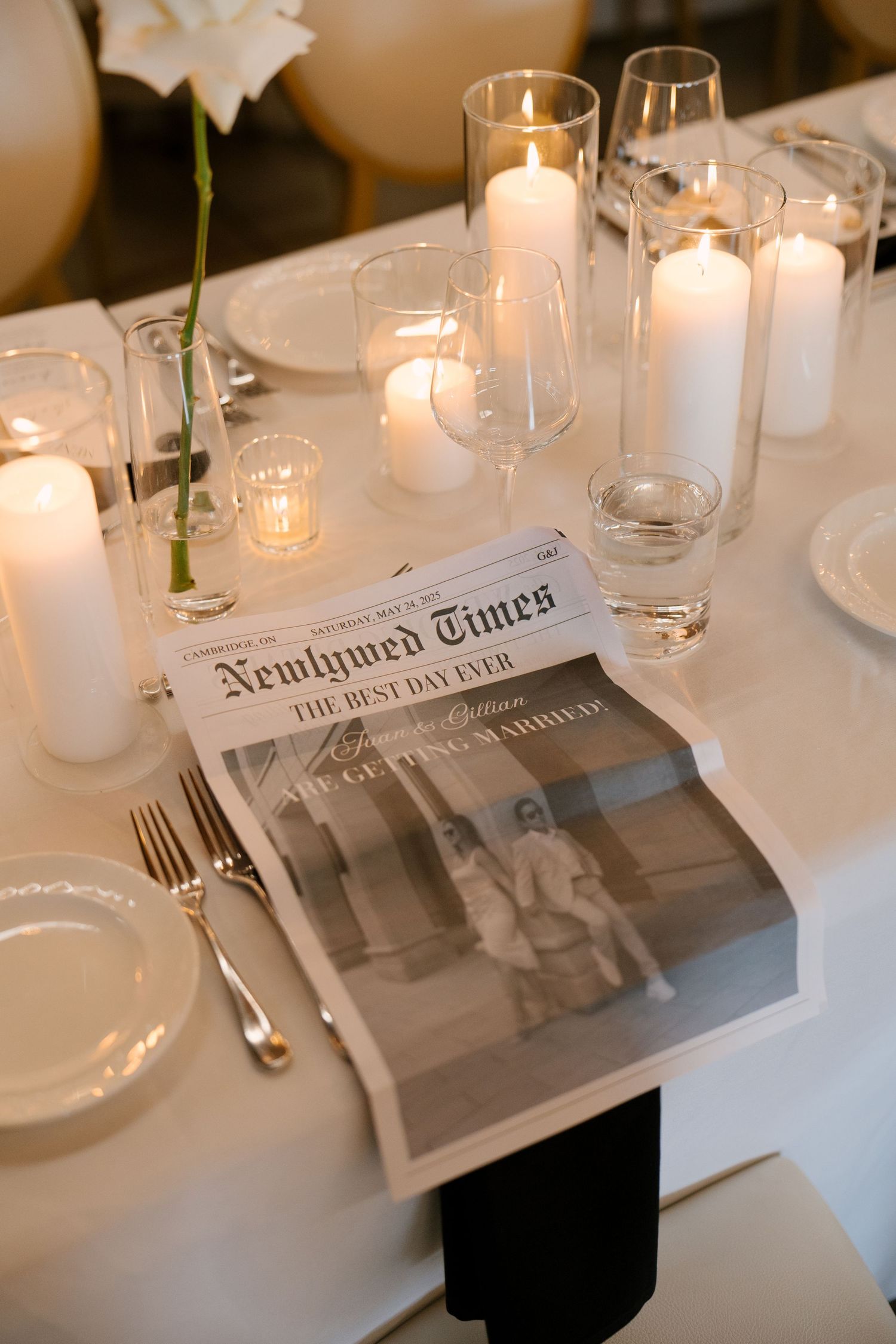 Wedding table place setting featuring a Newlywed Times newspaper program, white pillar candles, and elegant dinnerware at Tapestry Hall.