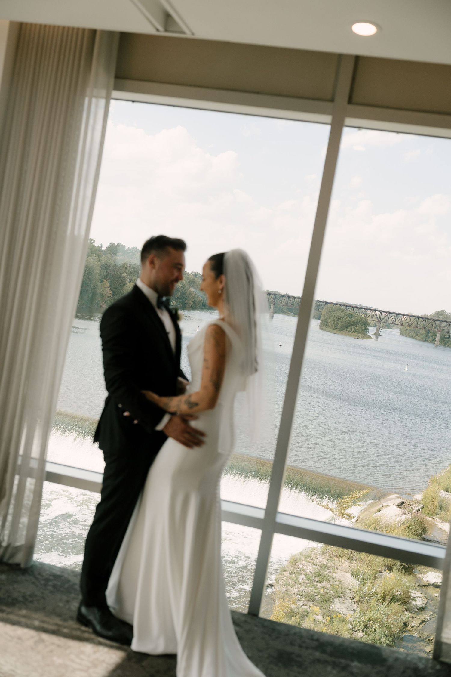 Bride and groom share a romantic moment by floor-to-ceiling windows overlooking a serene river on their wedding day at Cambridge Mill.