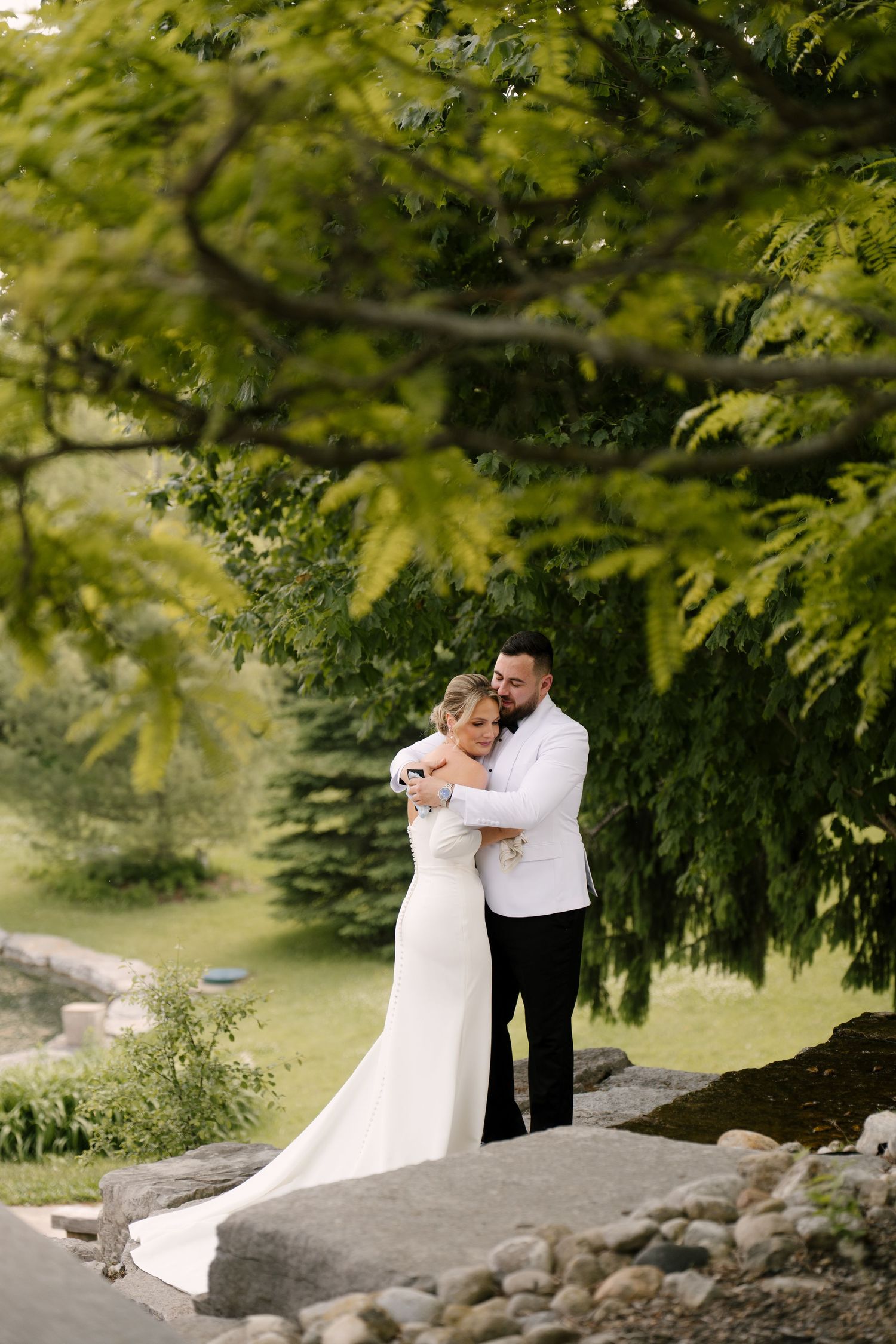 Bride and groom sharing a tender embrace under green trees during outdoor wedding portraits in a garden.