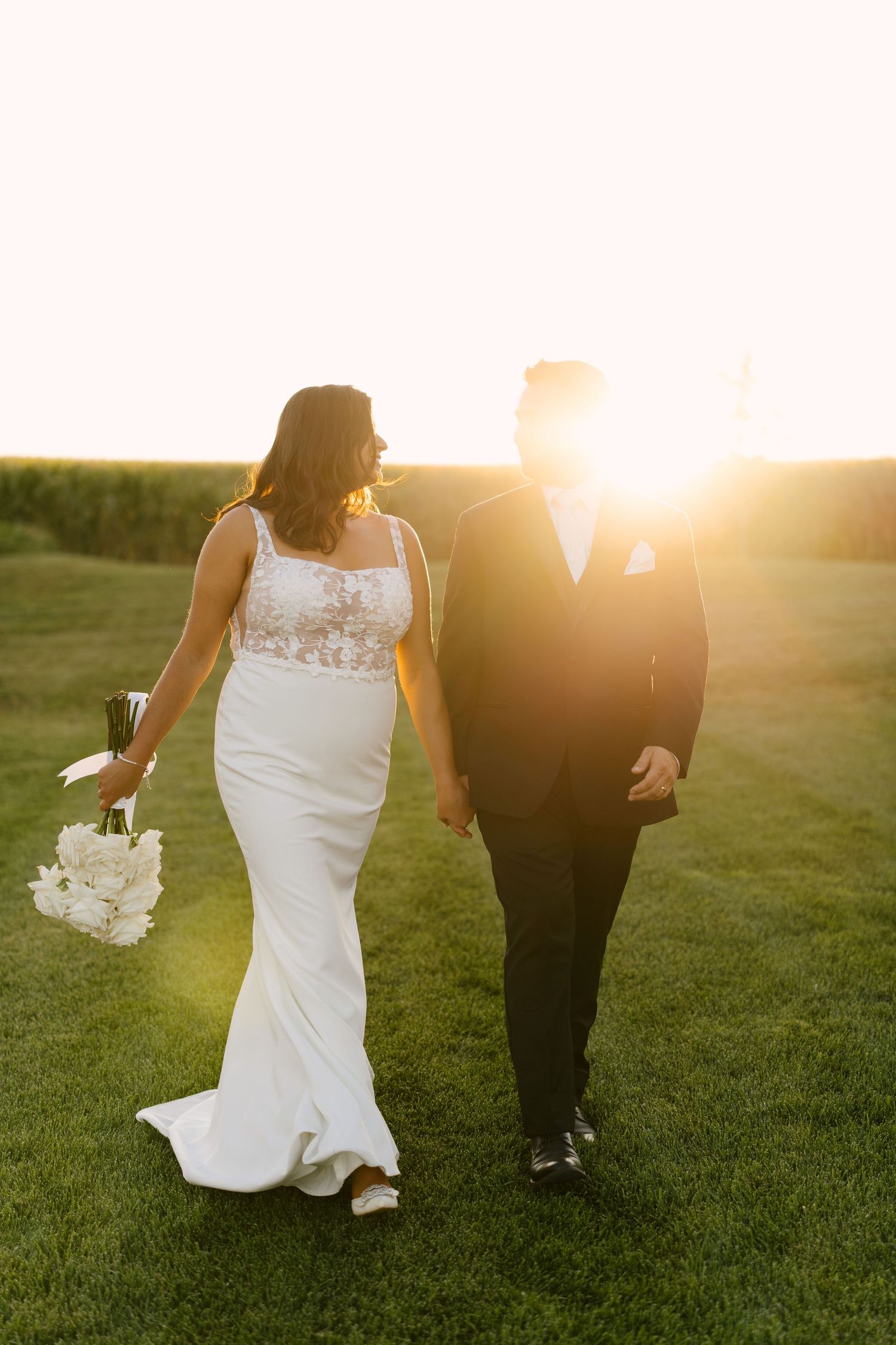 Bride and groom walking hand in hand on green grass at golden sunset, bride holding white bouquet in elegant fitted gown.