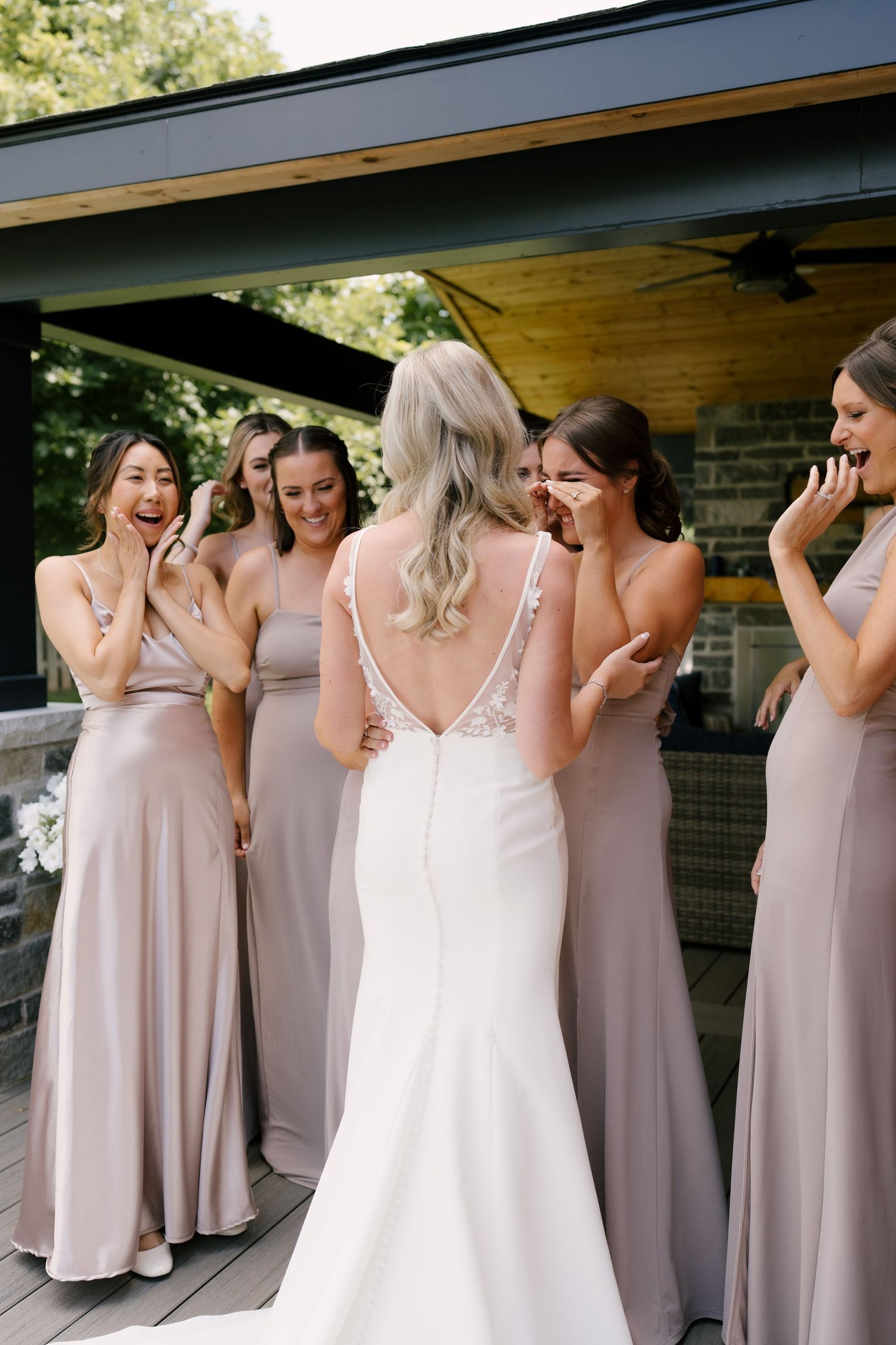 Bridesmaids in blush gowns reacting with joy as a blonde bride reveals her lace open-back wedding dress.